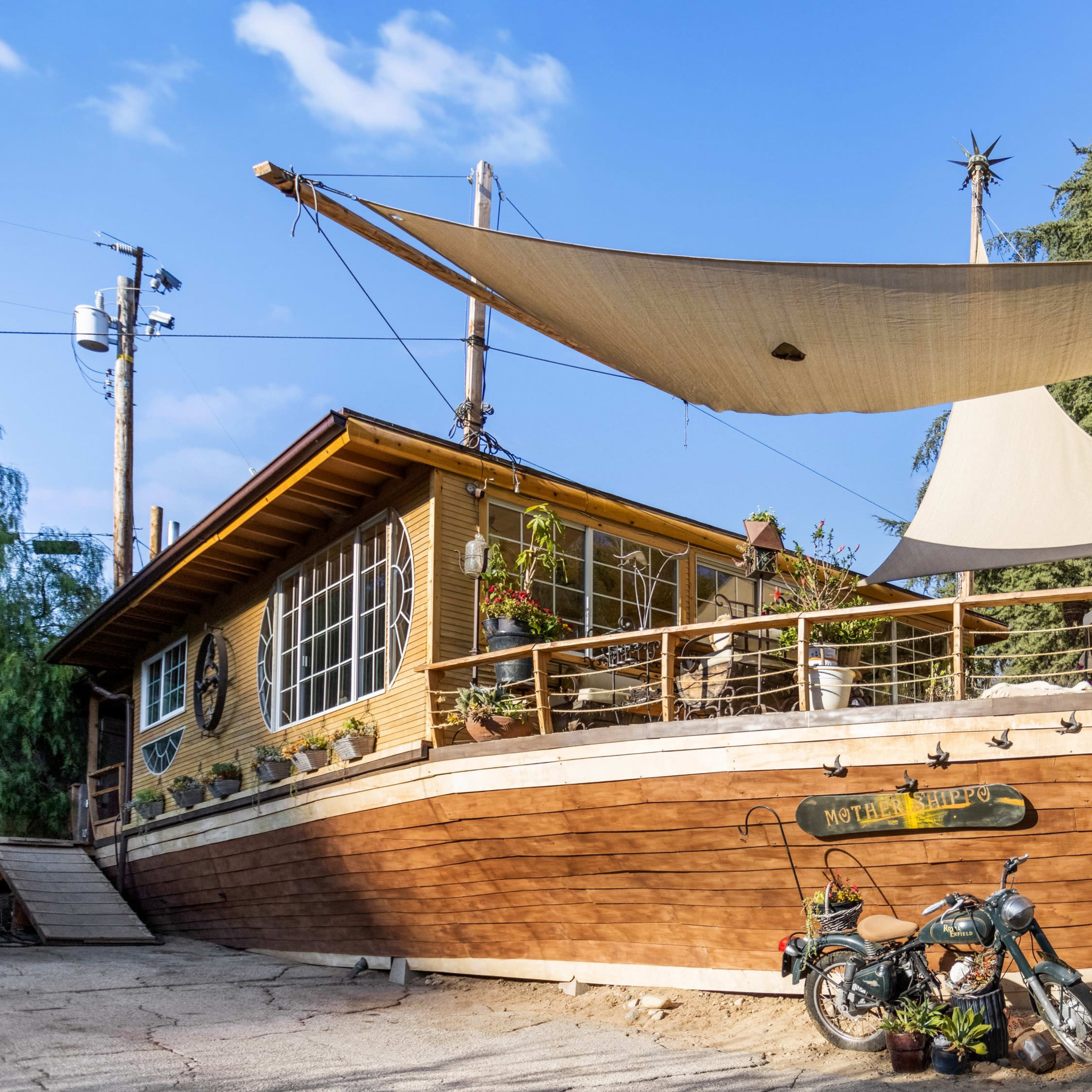 A wooden house designed to resemble a ship sits on a slope, with a large sail covering the deck and a motorcycle parked nearby.