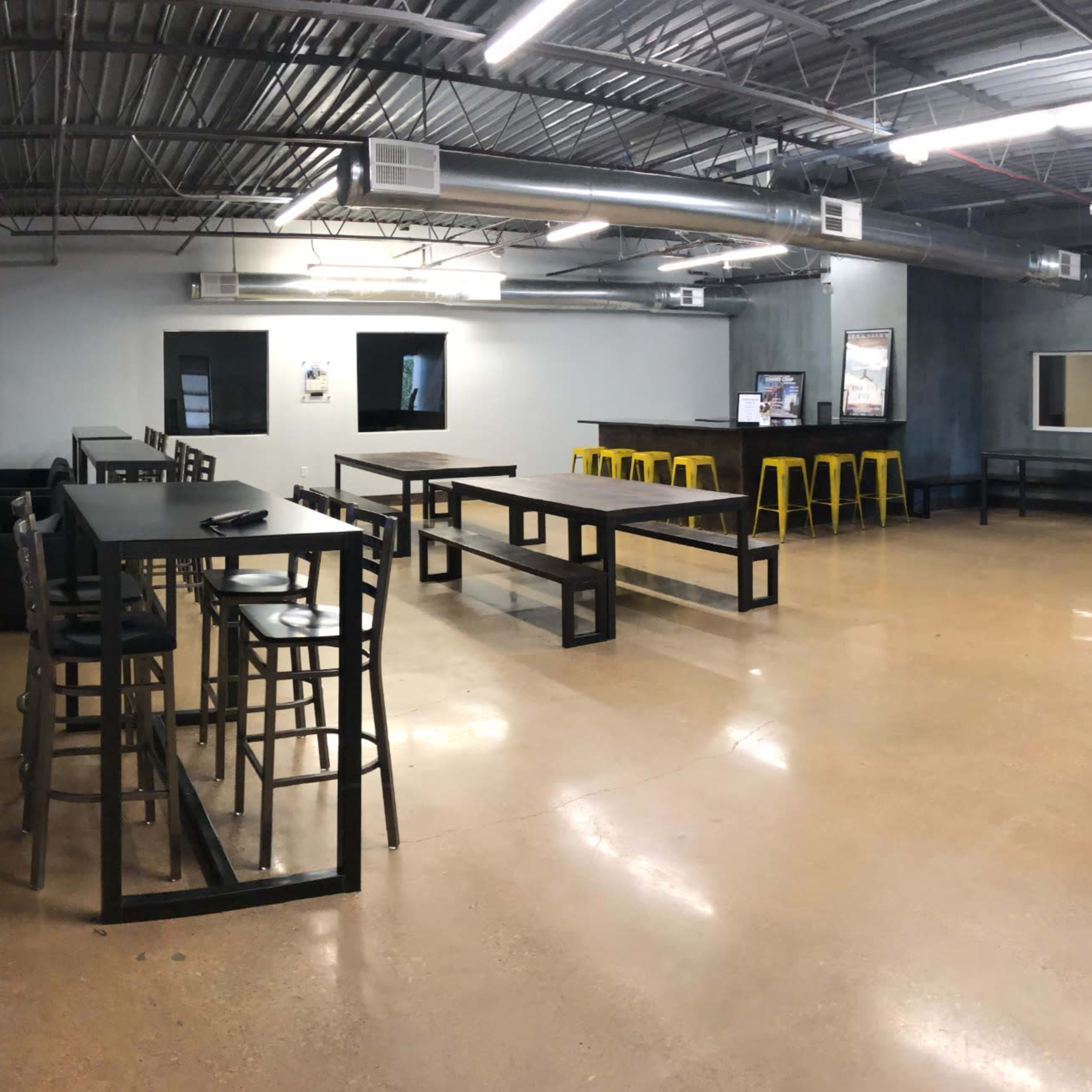 A spacious indoor area with tables, stools, and a couch, featuring an industrial-style design and multiple entry points.