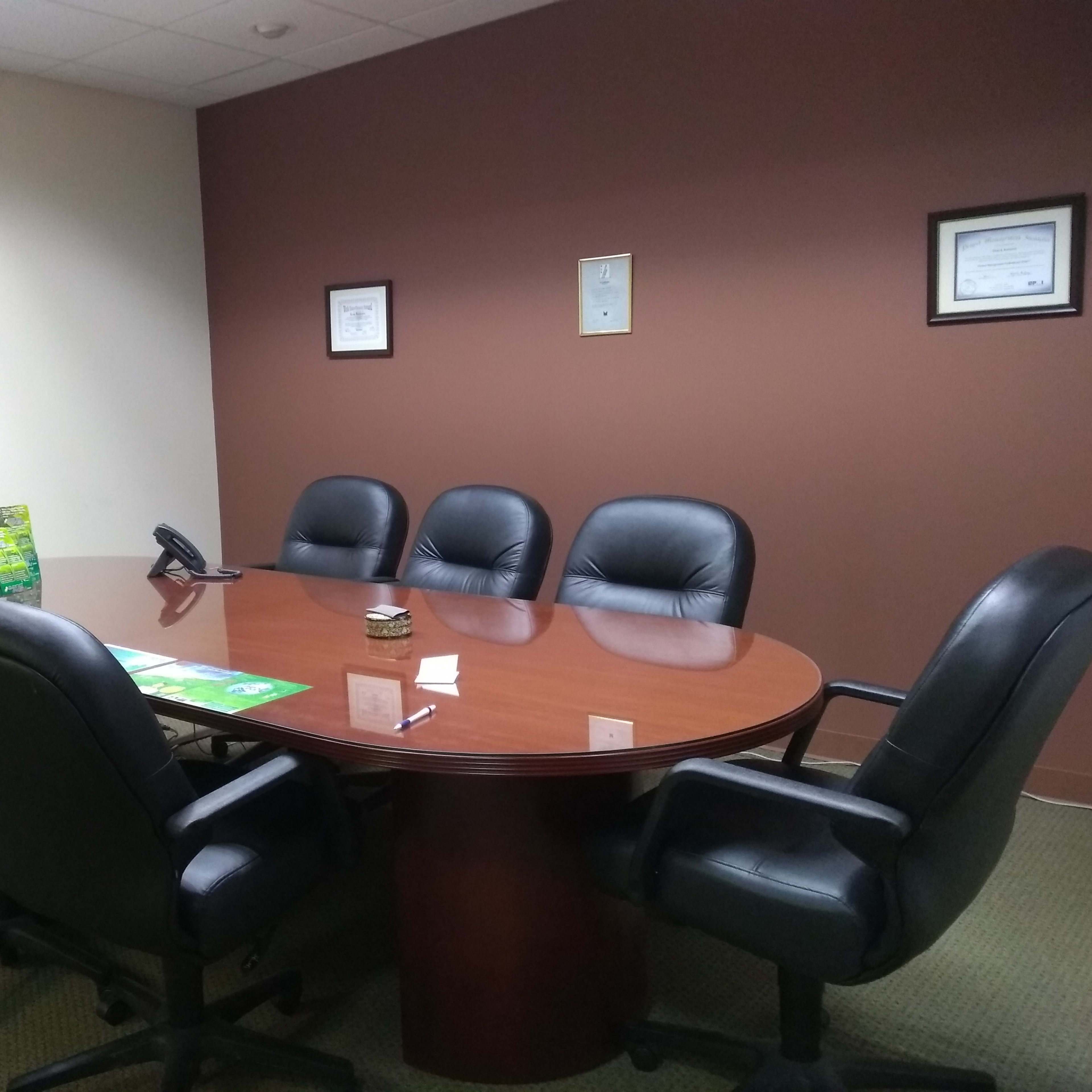 A conference room features a round wooden table surrounded by six black leather chairs, with a phone and materials on the table and certificates displayed on the wall.