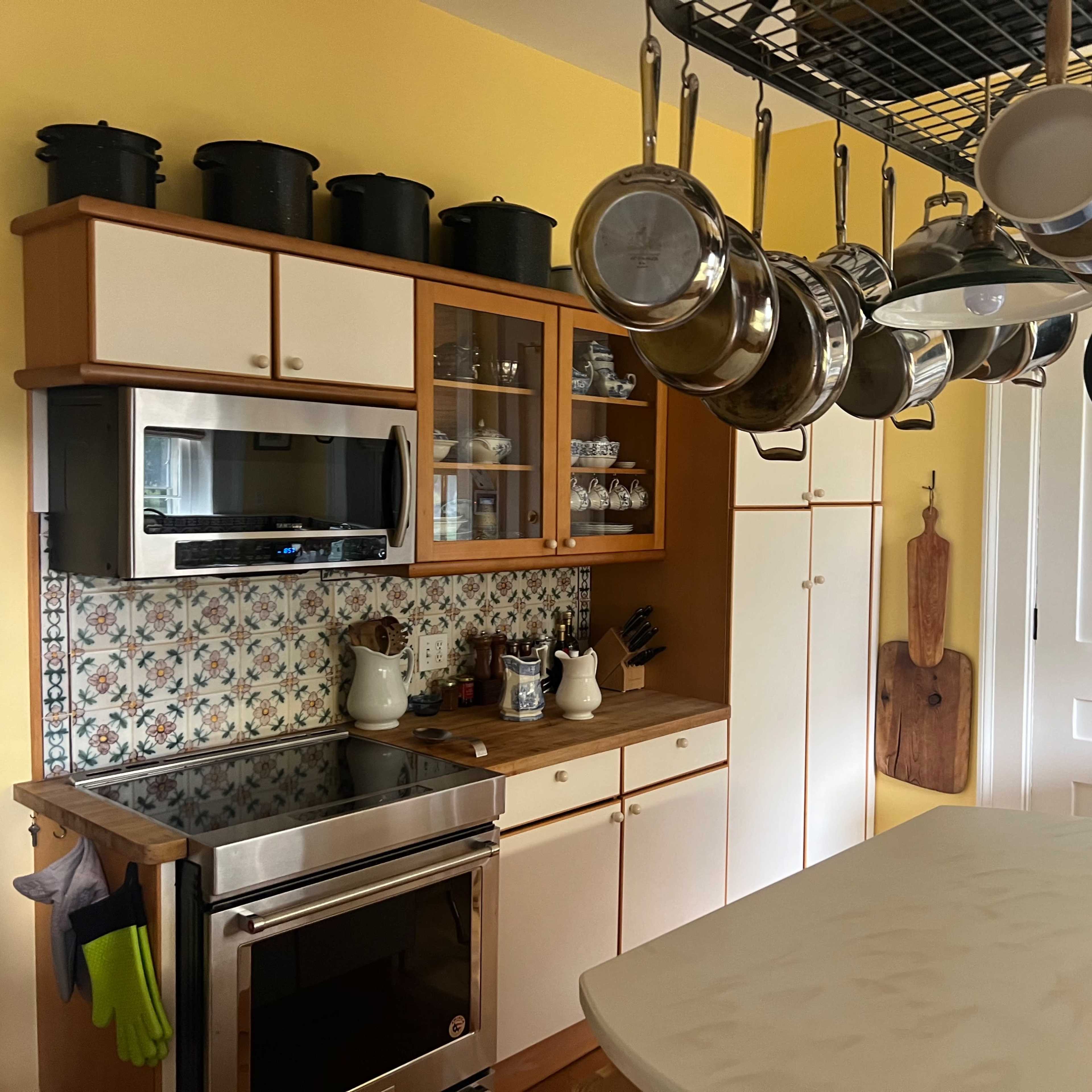 A kitchen with wooden cabinets, a countertop island, hanging pots and pans, and a tiled backsplash.