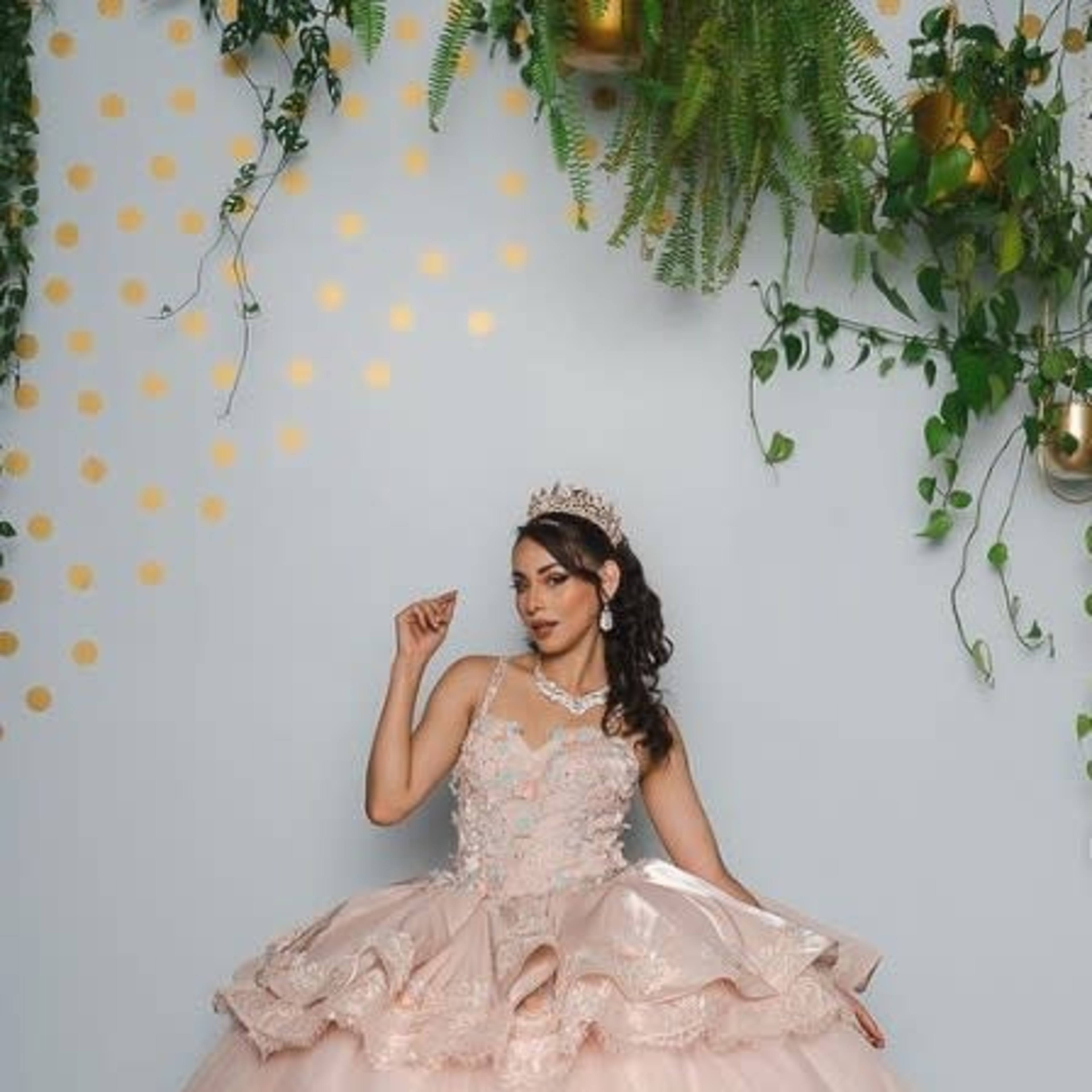 A woman in a large, ornate pink dress poses against a light blue wall adorned with greenery and golden polka dots.