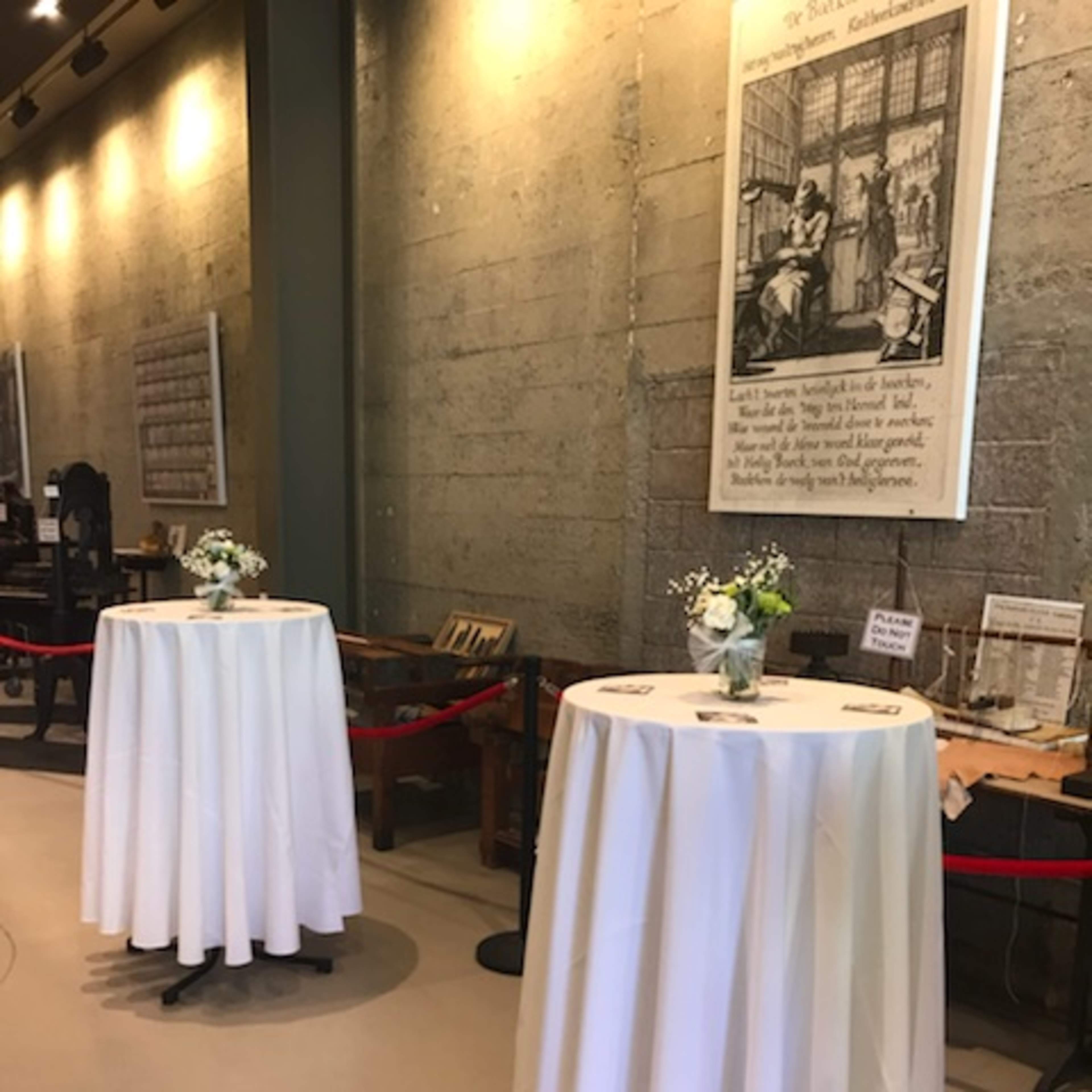 Two round tables covered with white tablecloths, set up in a well-lit space with a concrete wall and a large framed artwork in the background.