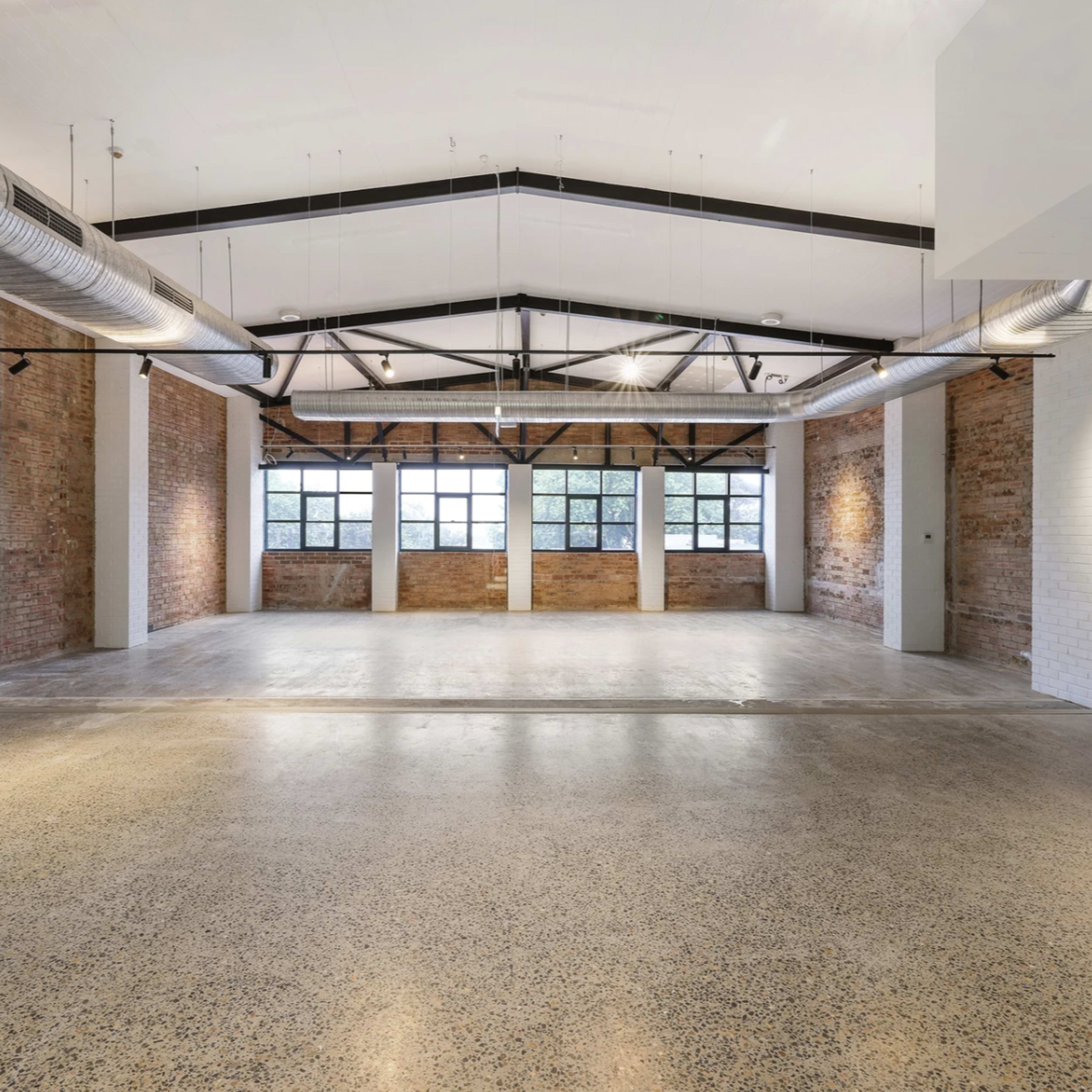 A spacious, empty industrial-style room with exposed brick walls, concrete flooring, and large windows offering natural light.