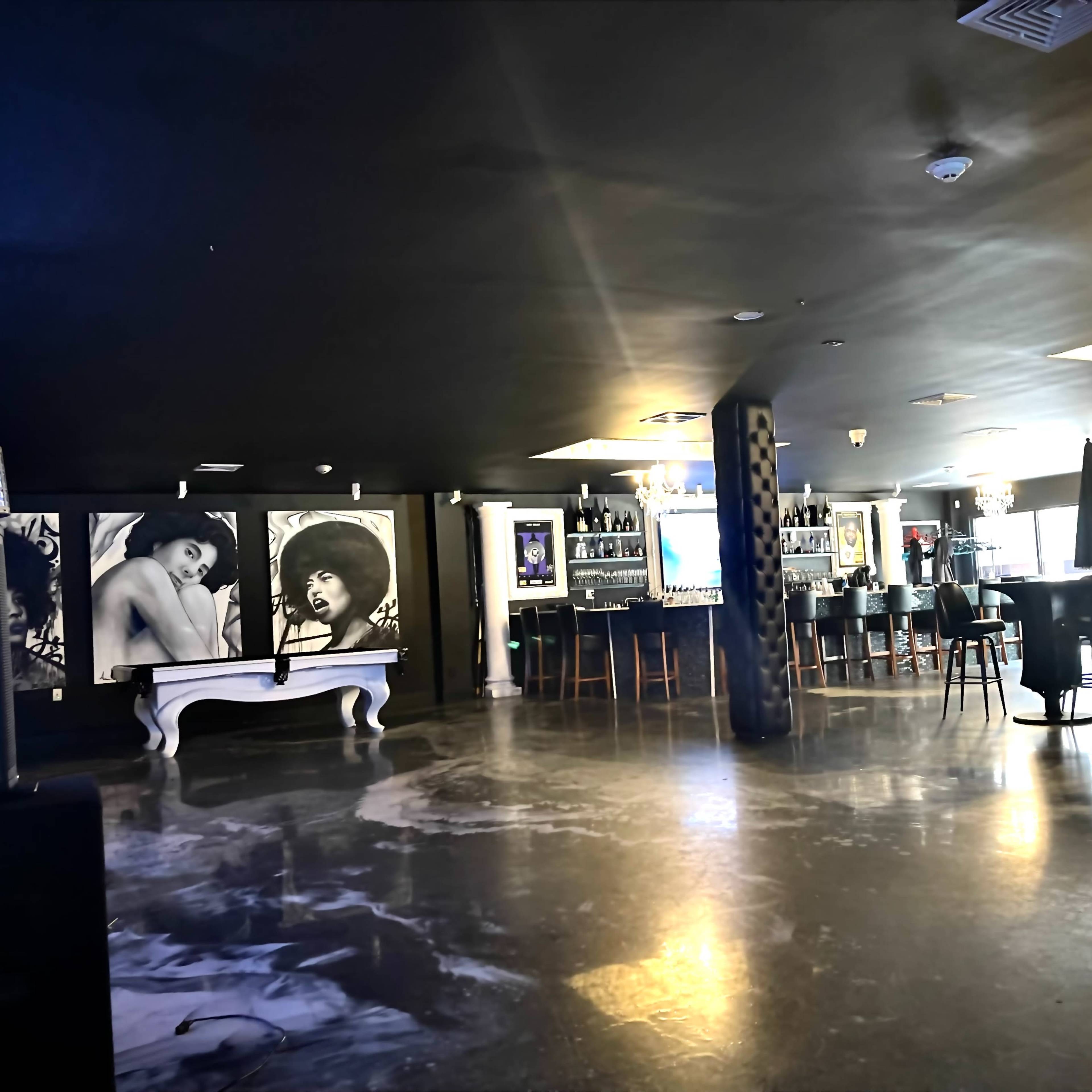 The interior of a dimly lit bar features large black-and-white portraits on the walls and a long counter with stools.
