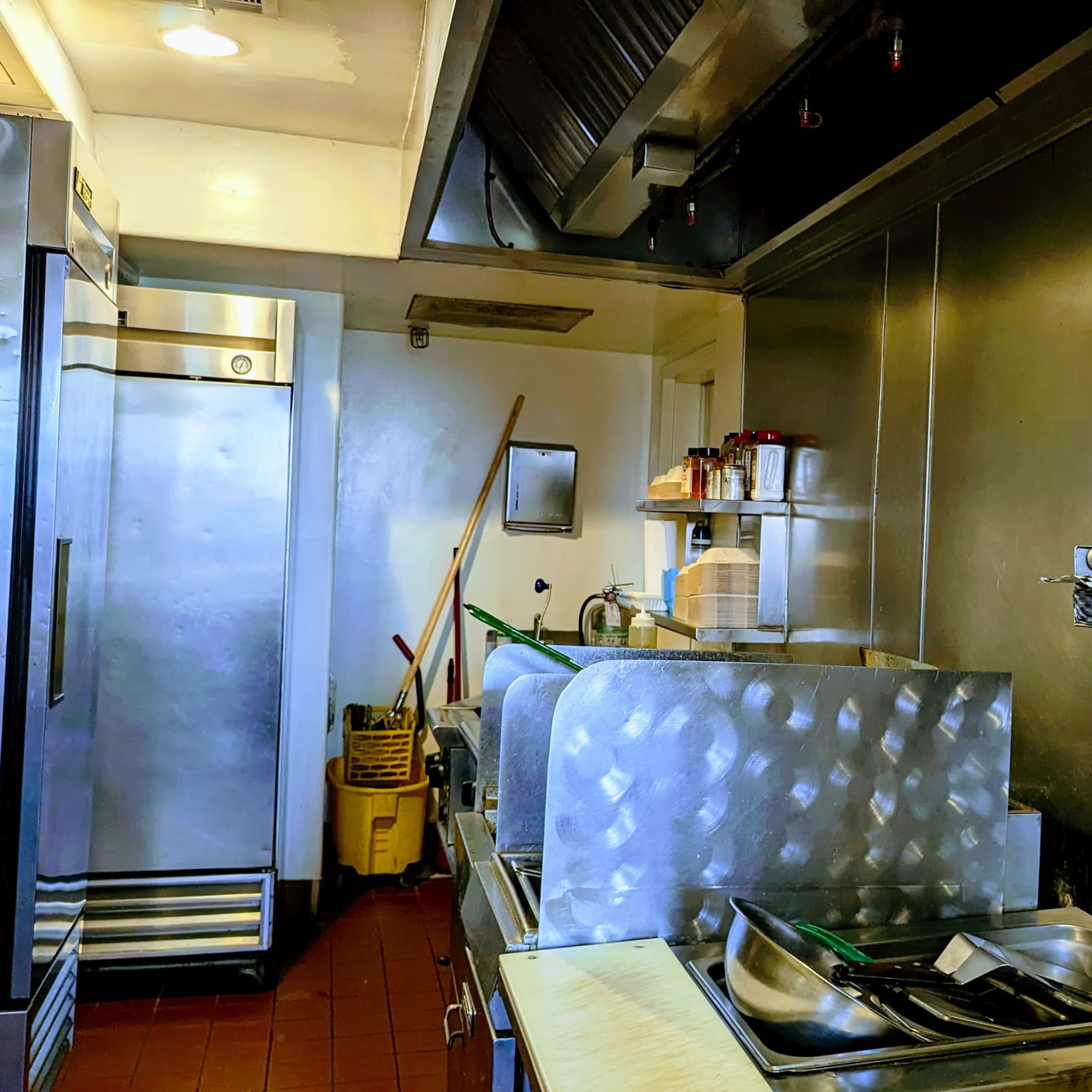 A commercial kitchen with stainless steel surfaces, a double sink, shelves holding dishes, and a large refrigerator.