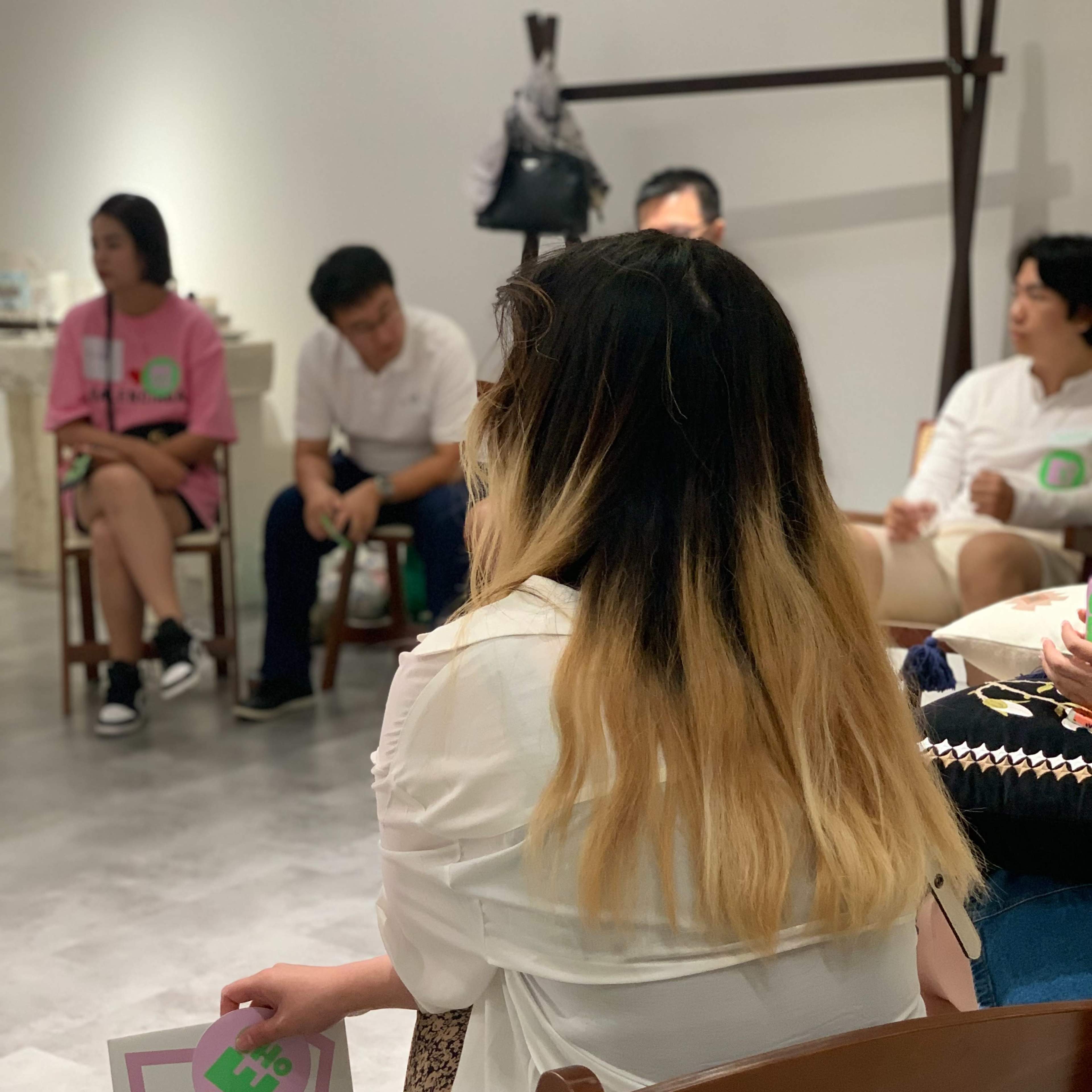 A woman with long, ombre hair sits in a chair holding a sign while other people are gathered around her in a room.
