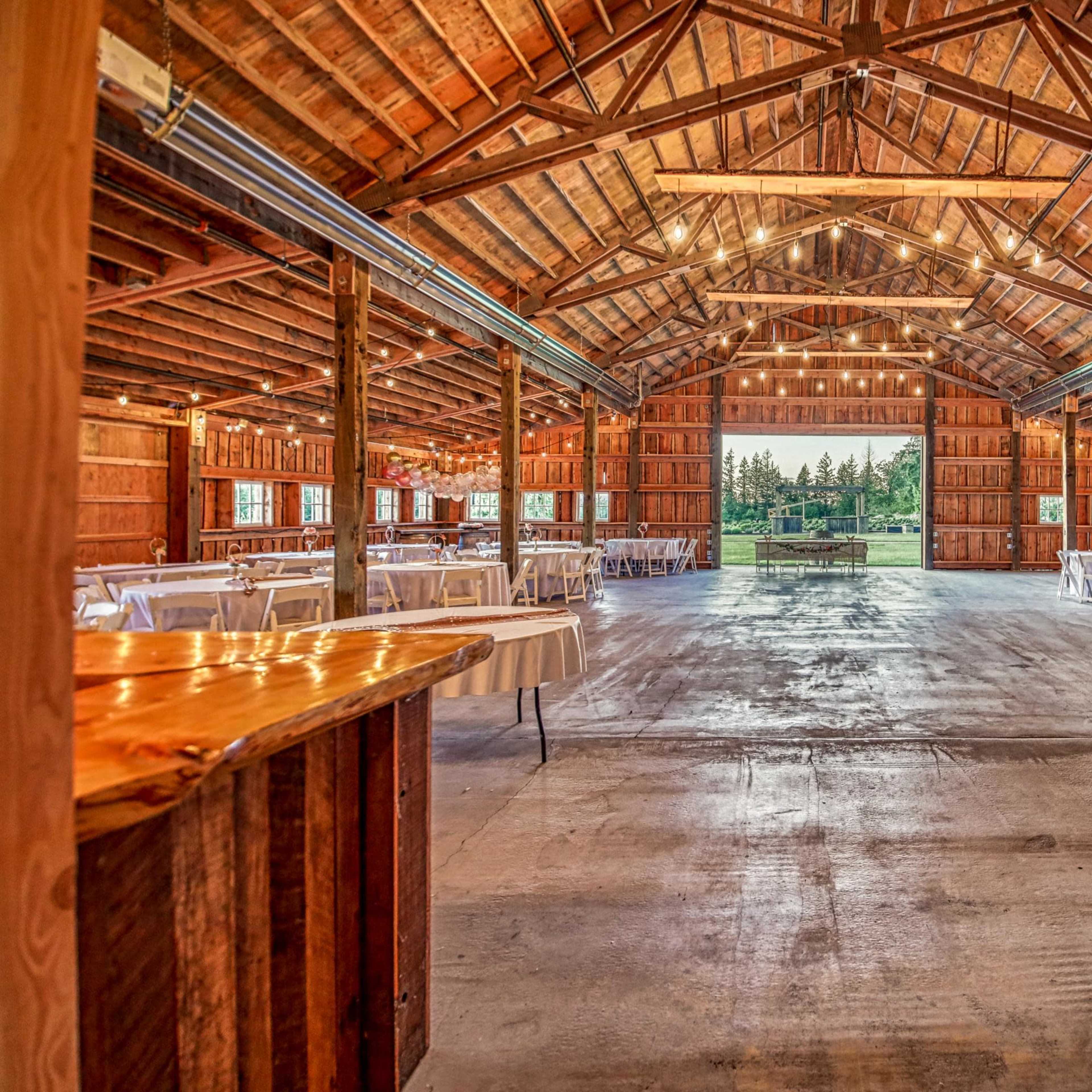 The interior of a spacious barn venue features wooden beams, table setups, and large openings that lead to an outdoor view.