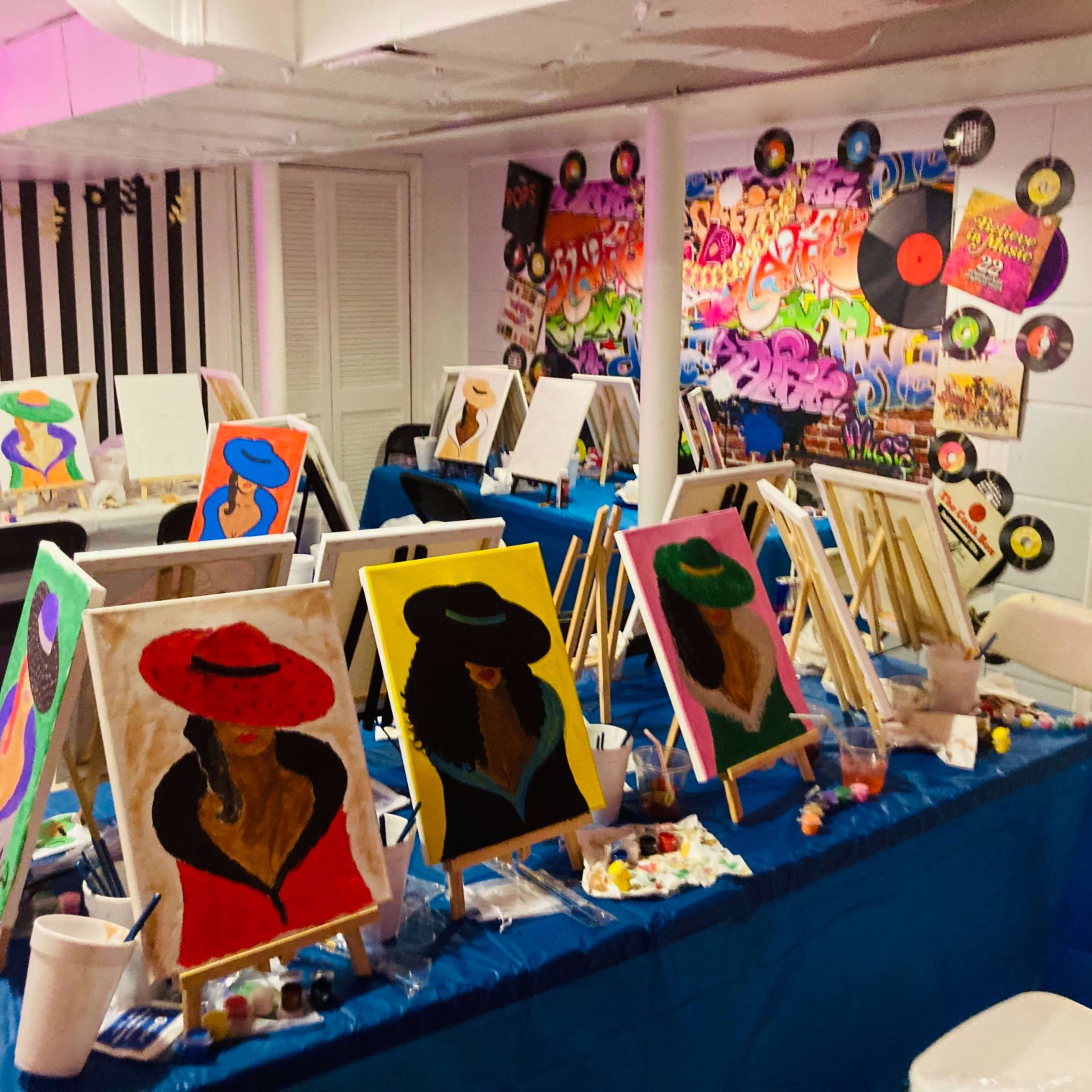 A brightly decorated art studio with multiple easels displaying paintings of women wearing hats, surrounded by painting supplies and a colorful graffiti-style mural on the wall.