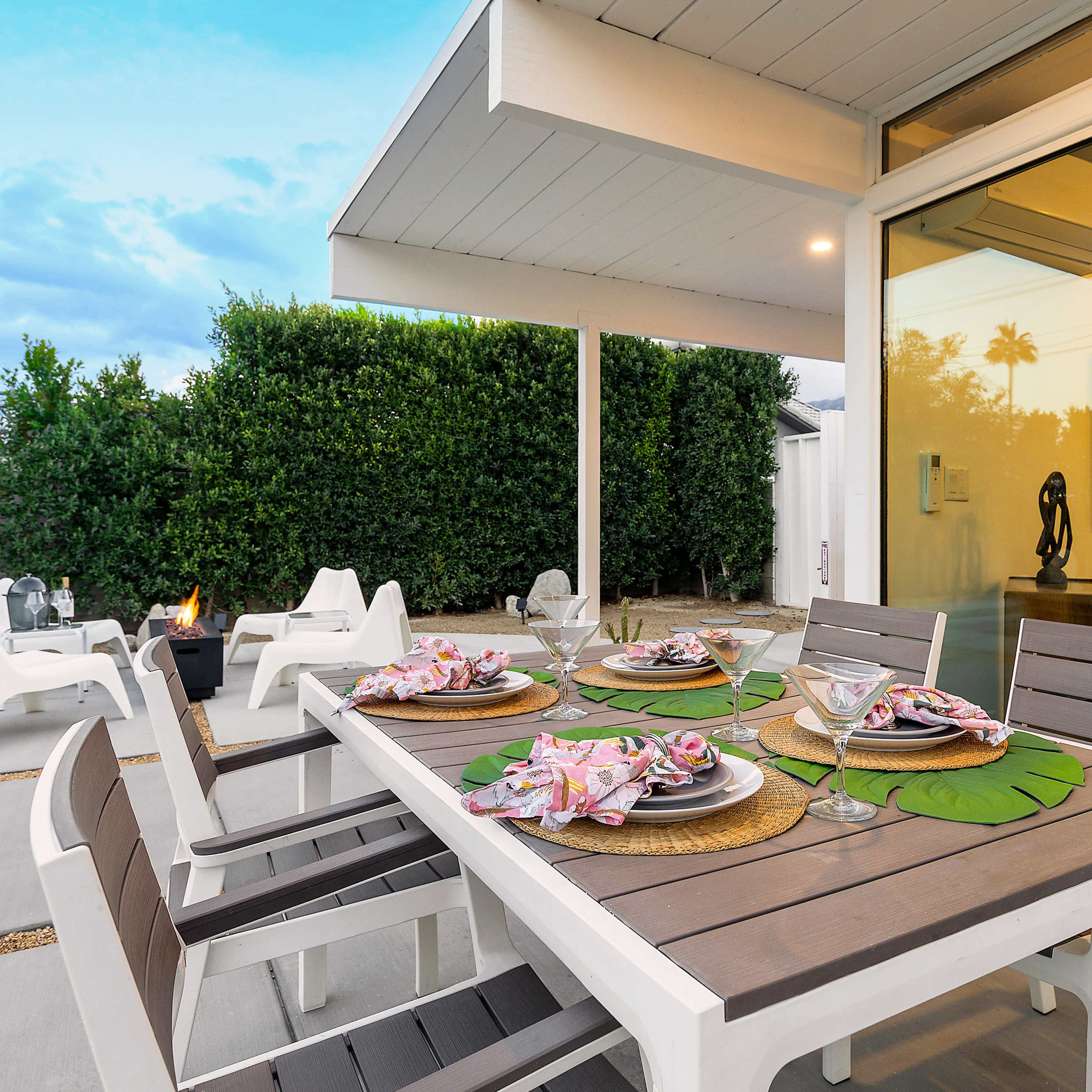 A modern outdoor dining area with a table set for four, featuring plates and glassware, and a view of a landscaped garden in the background.
