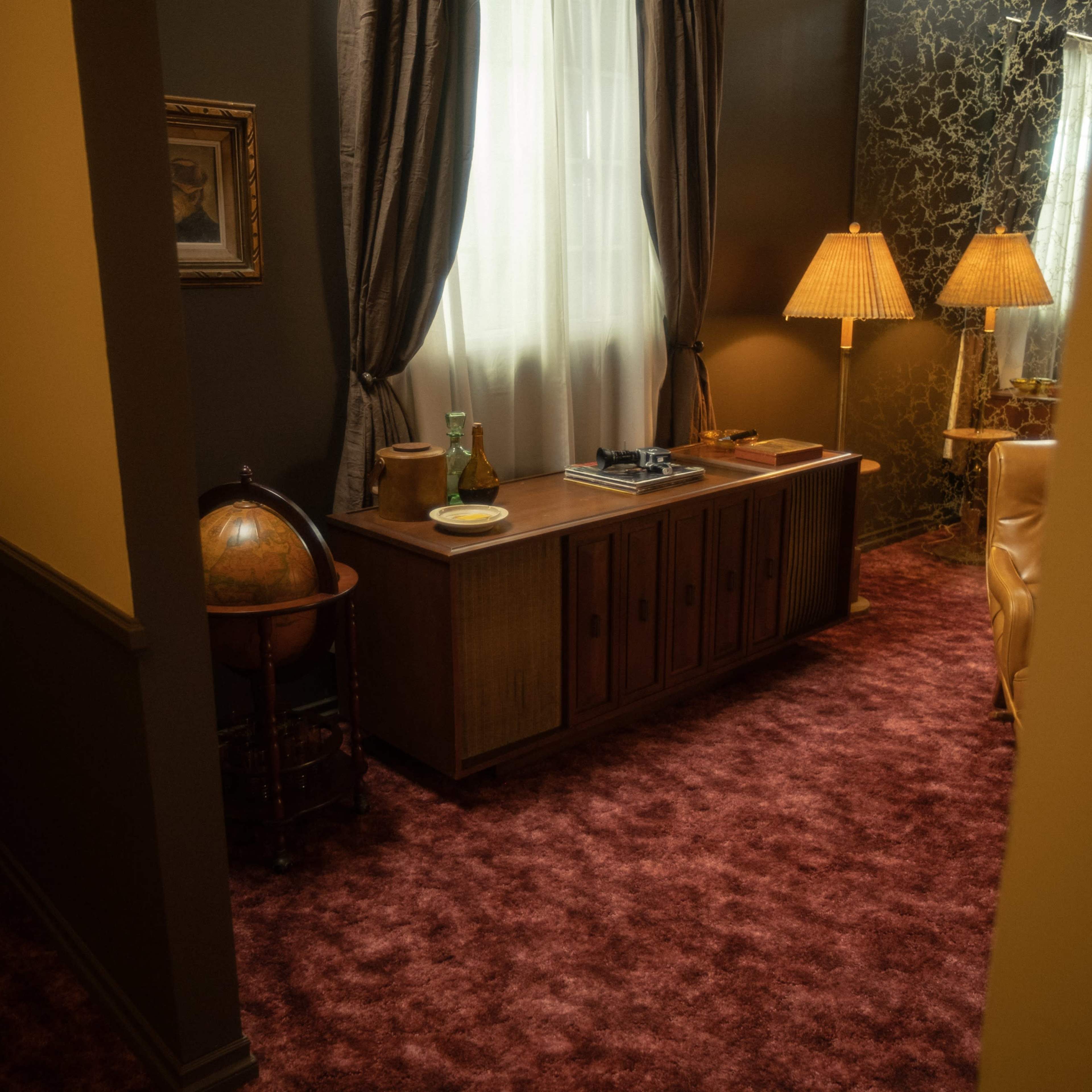 A room with a brown carpet, a wooden sideboard, a globe, and two lamps, illuminated by natural light from a window.