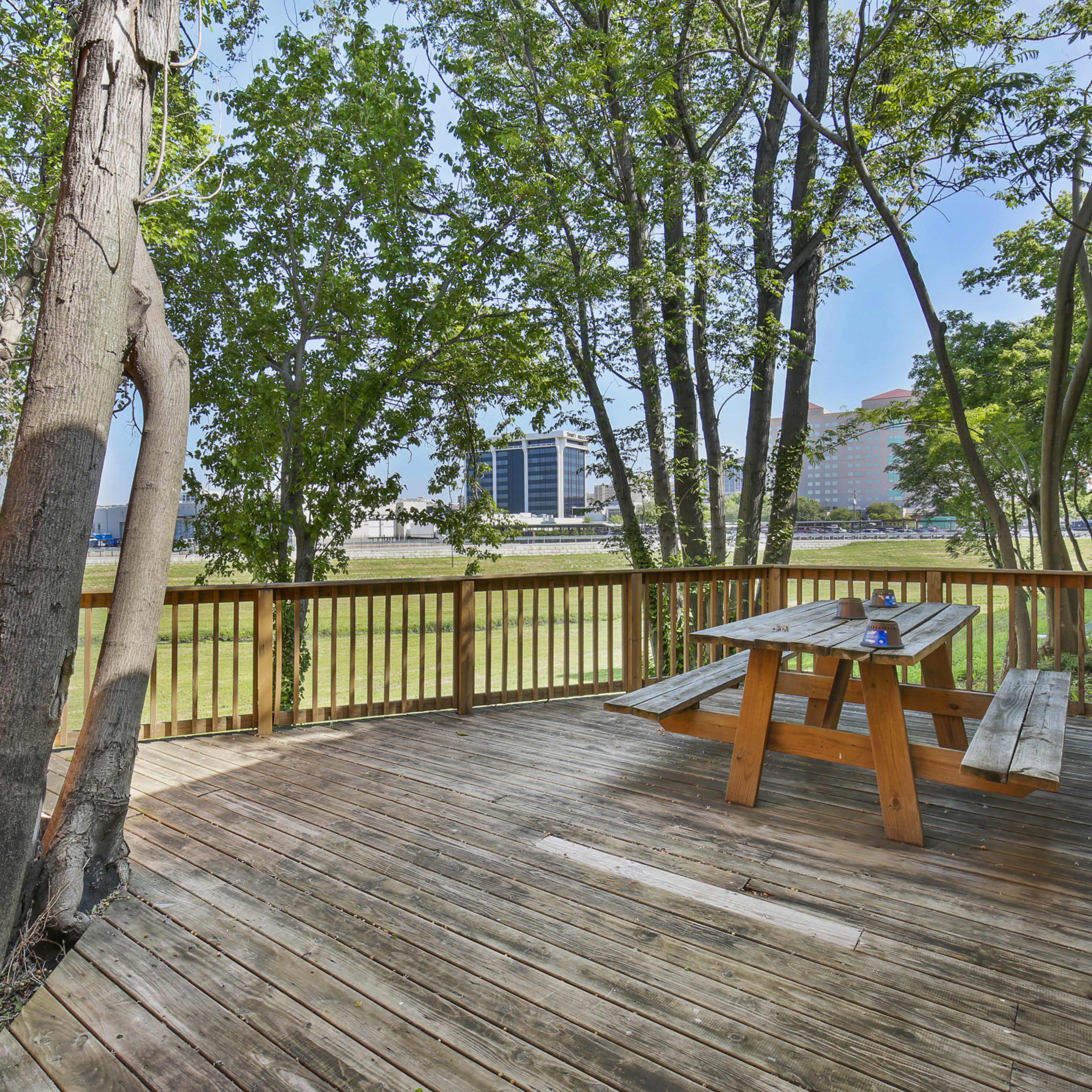 A wooden deck with a picnic table is surrounded by trees and overlooks a grassy area and buildings in the distance.