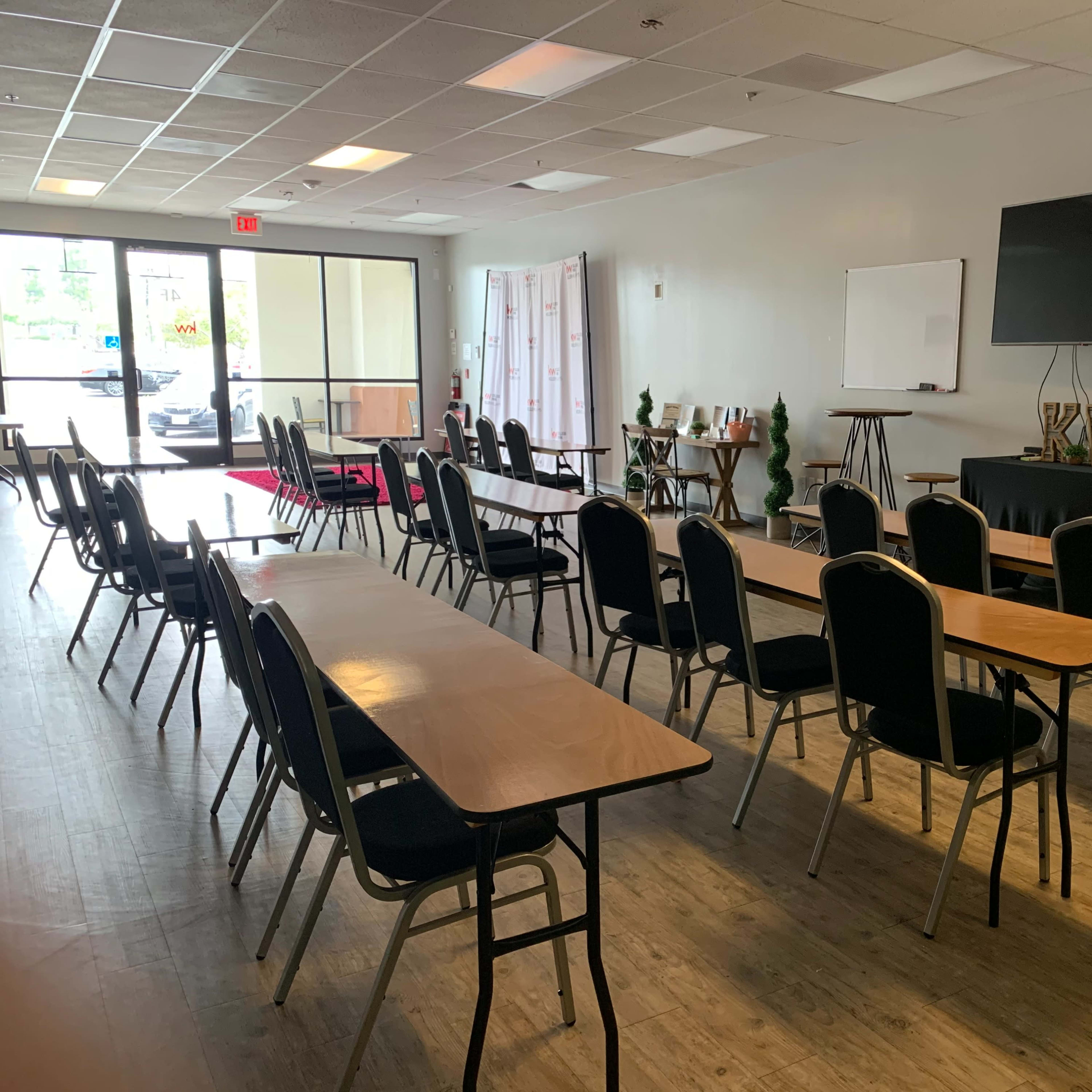 An interior room set up with multiple rows of tables and chairs, a large television, and a whiteboard at the front.