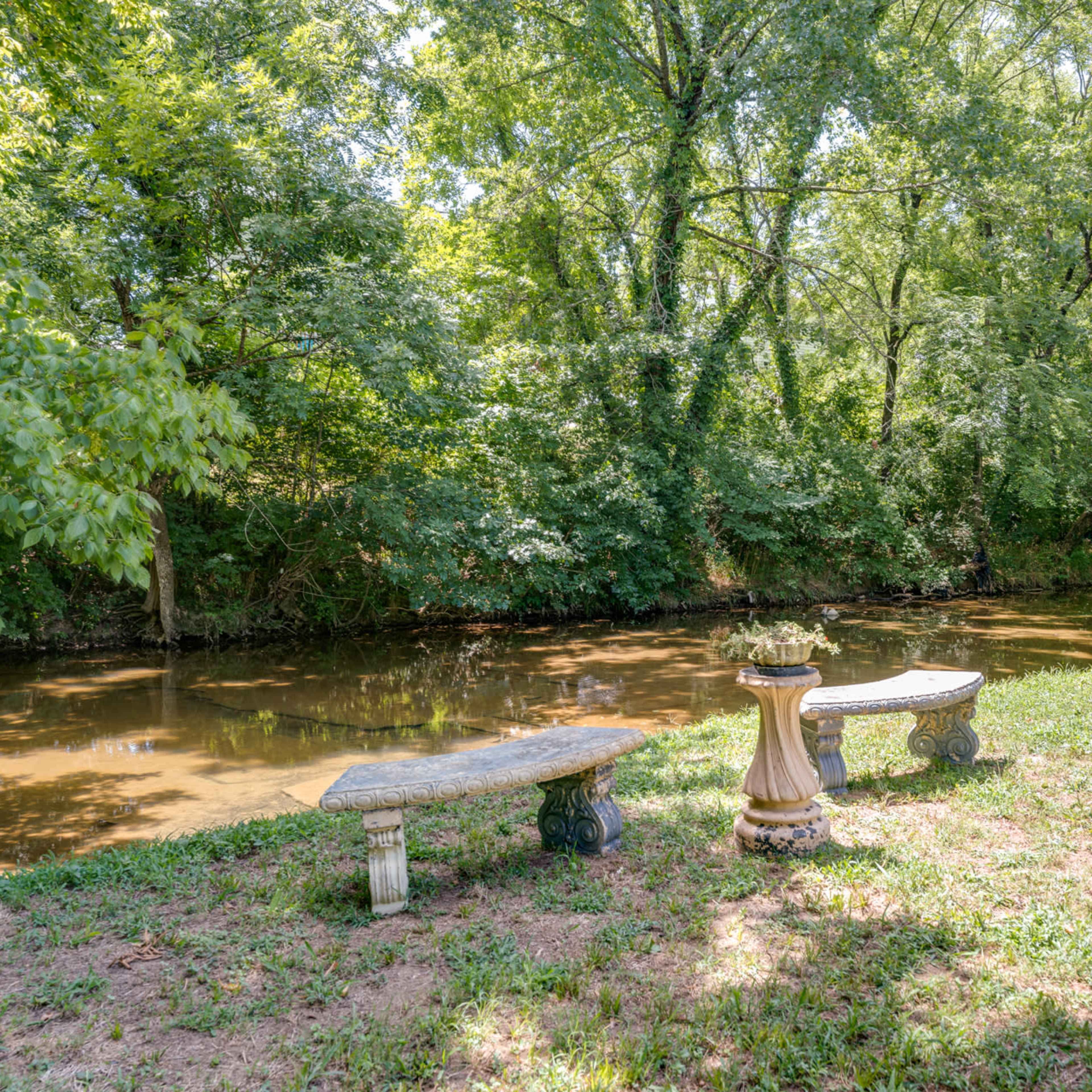 Two stone benches positioned along a grassy area beside a calm, tree-lined creek.