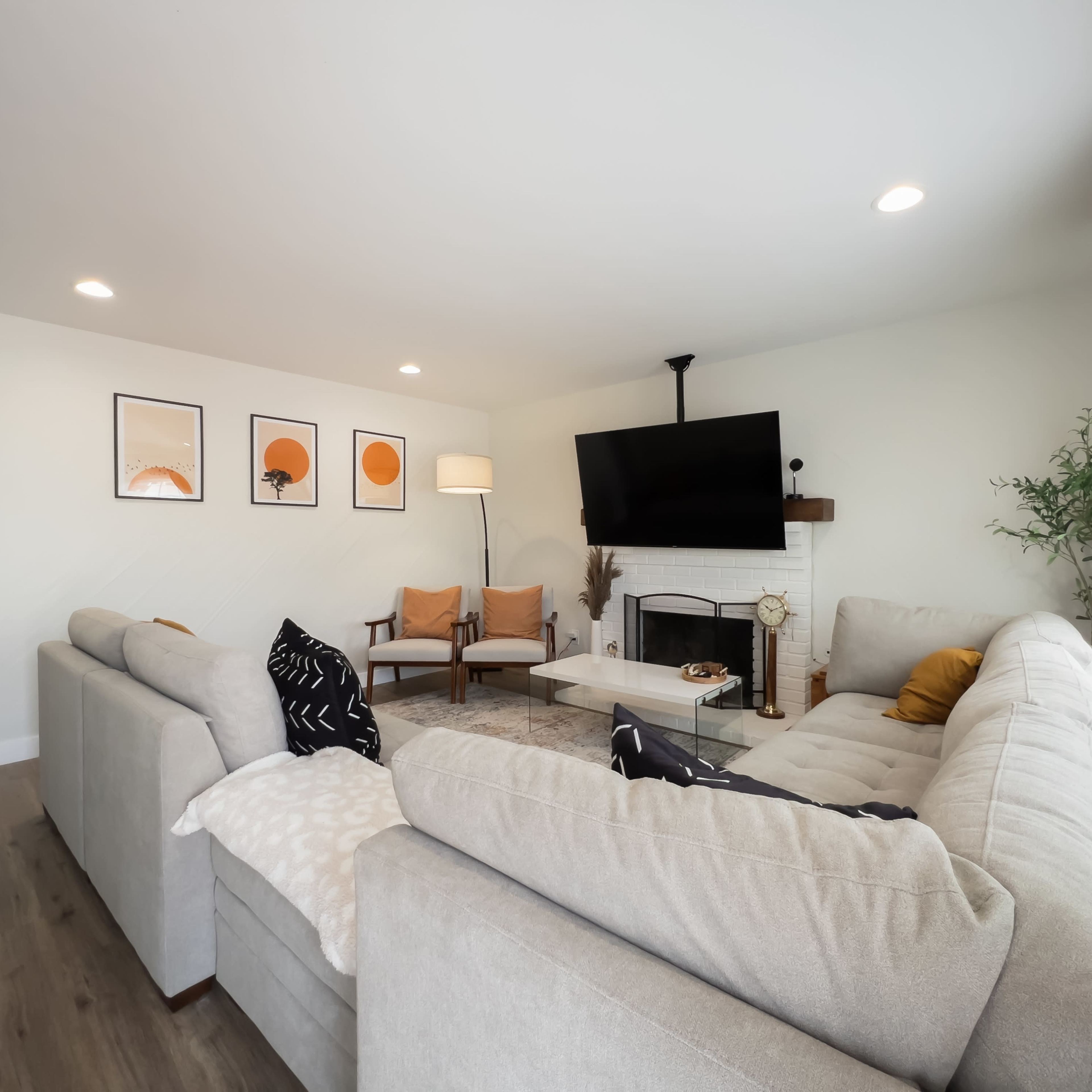 A modern living room features a large sectional sofa, a coffee table, and artwork on the walls, with a TV mounted above a fireplace.