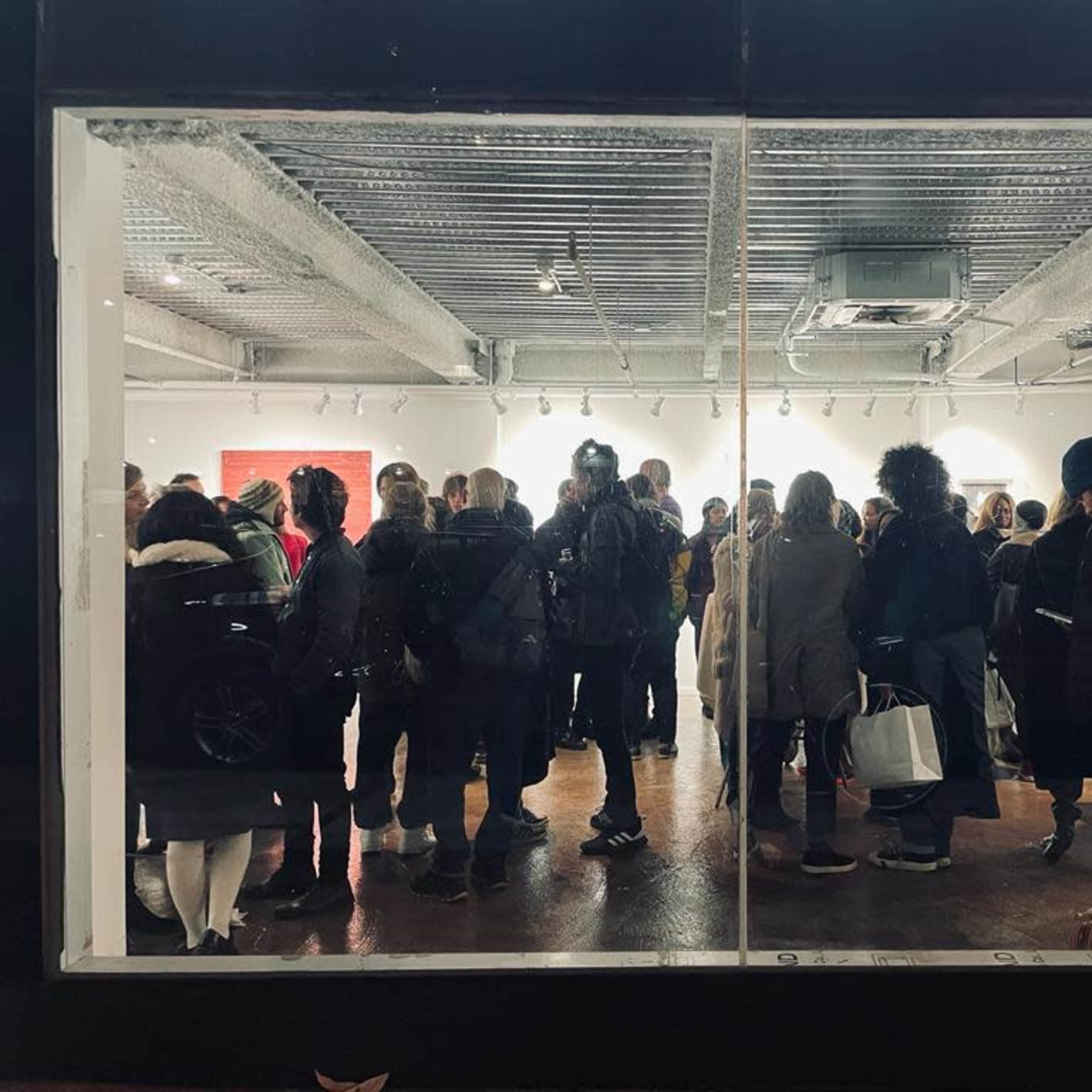 A crowd gathers inside an art gallery, visible through large windows, during an exhibition opening.