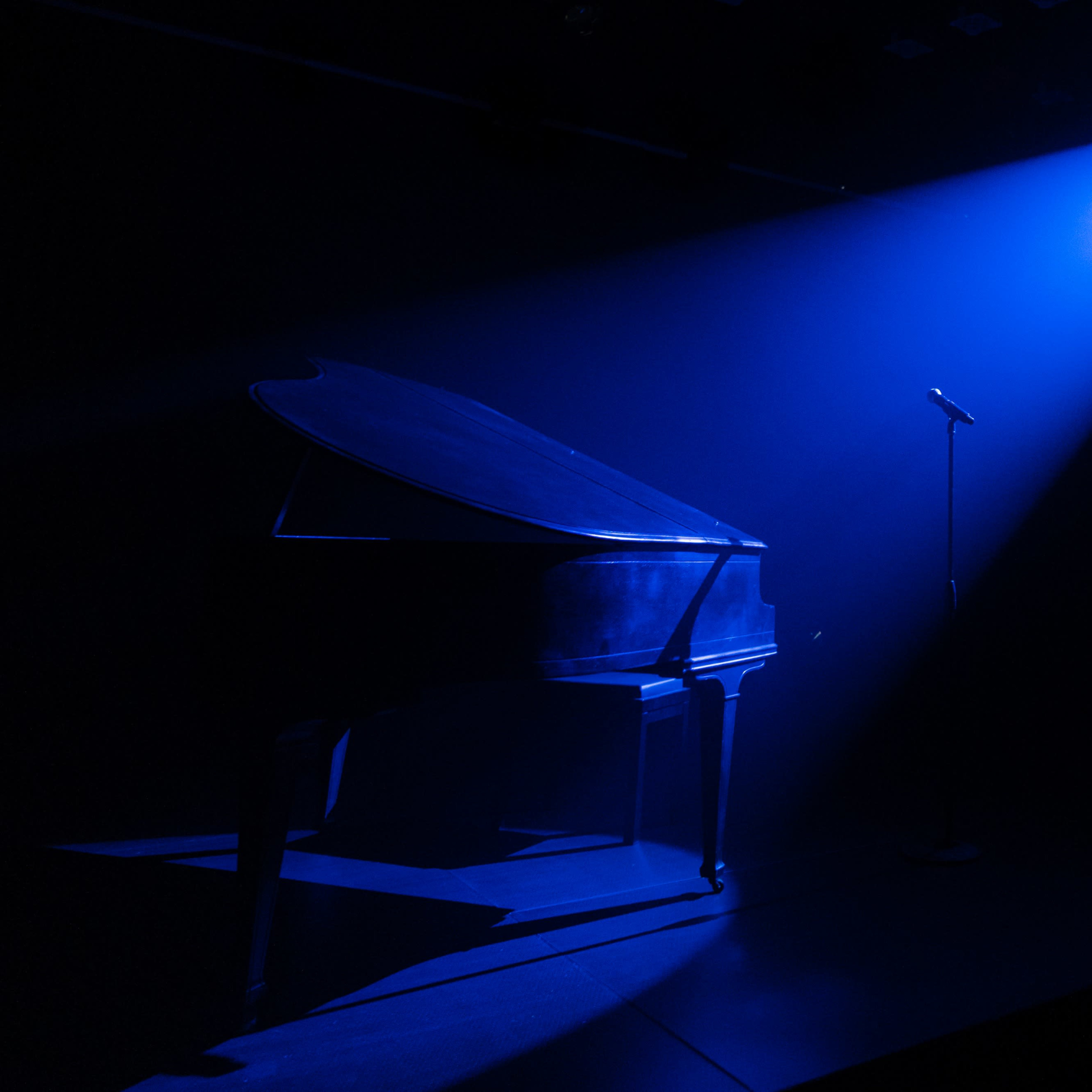 A grand piano is illuminated by a blue spotlight, with a microphone positioned nearby in a dark setting.
