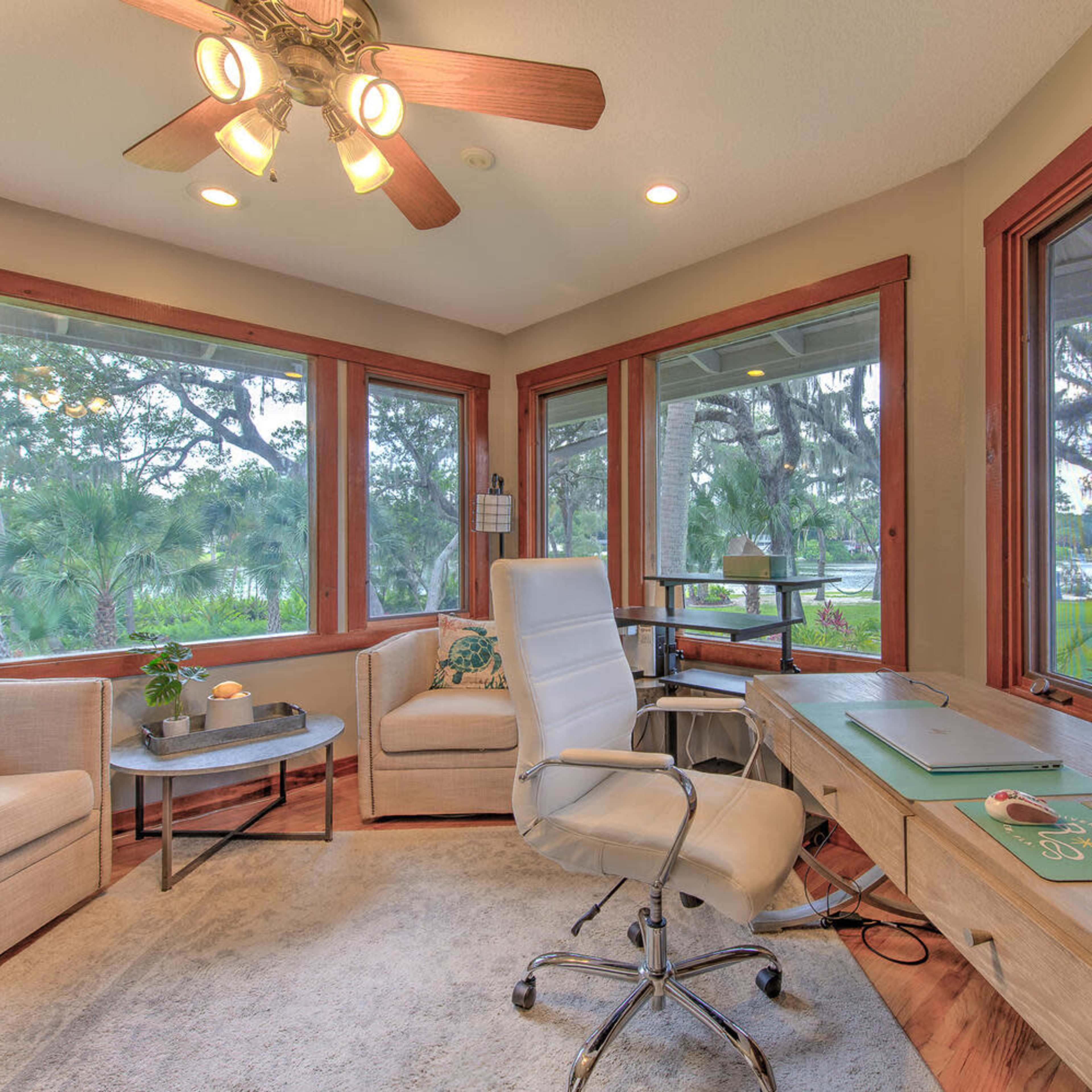 A well-lit home office features a desk, swivel chair, and comfortable seating arranged by large windows overlooking greenery.