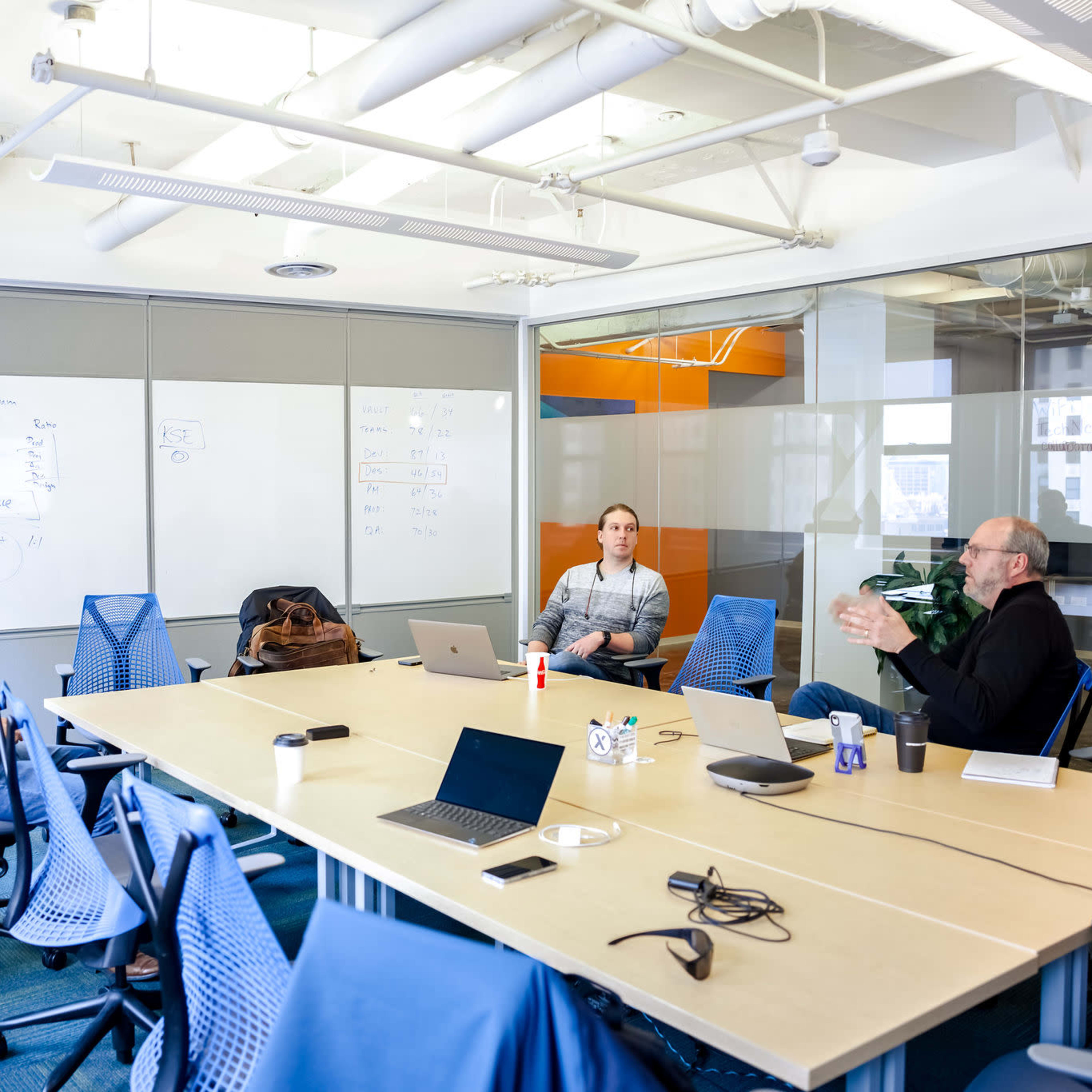 Three people are seated around a conference table in a bright meeting room with a whiteboard and laptops.