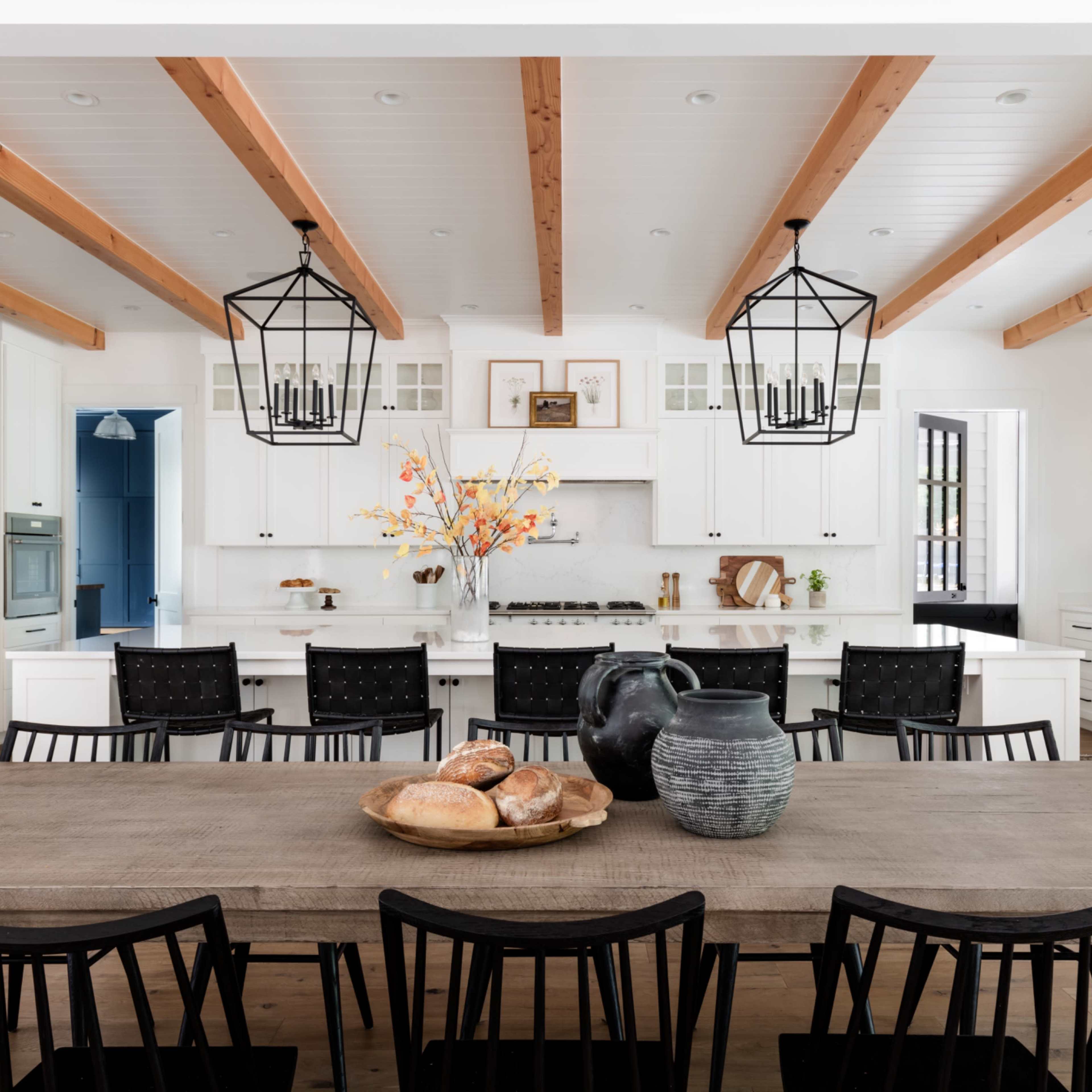 A spacious kitchen features a long wooden dining table surrounded by black chairs, with white cabinetry and exposed wooden beams overhead.