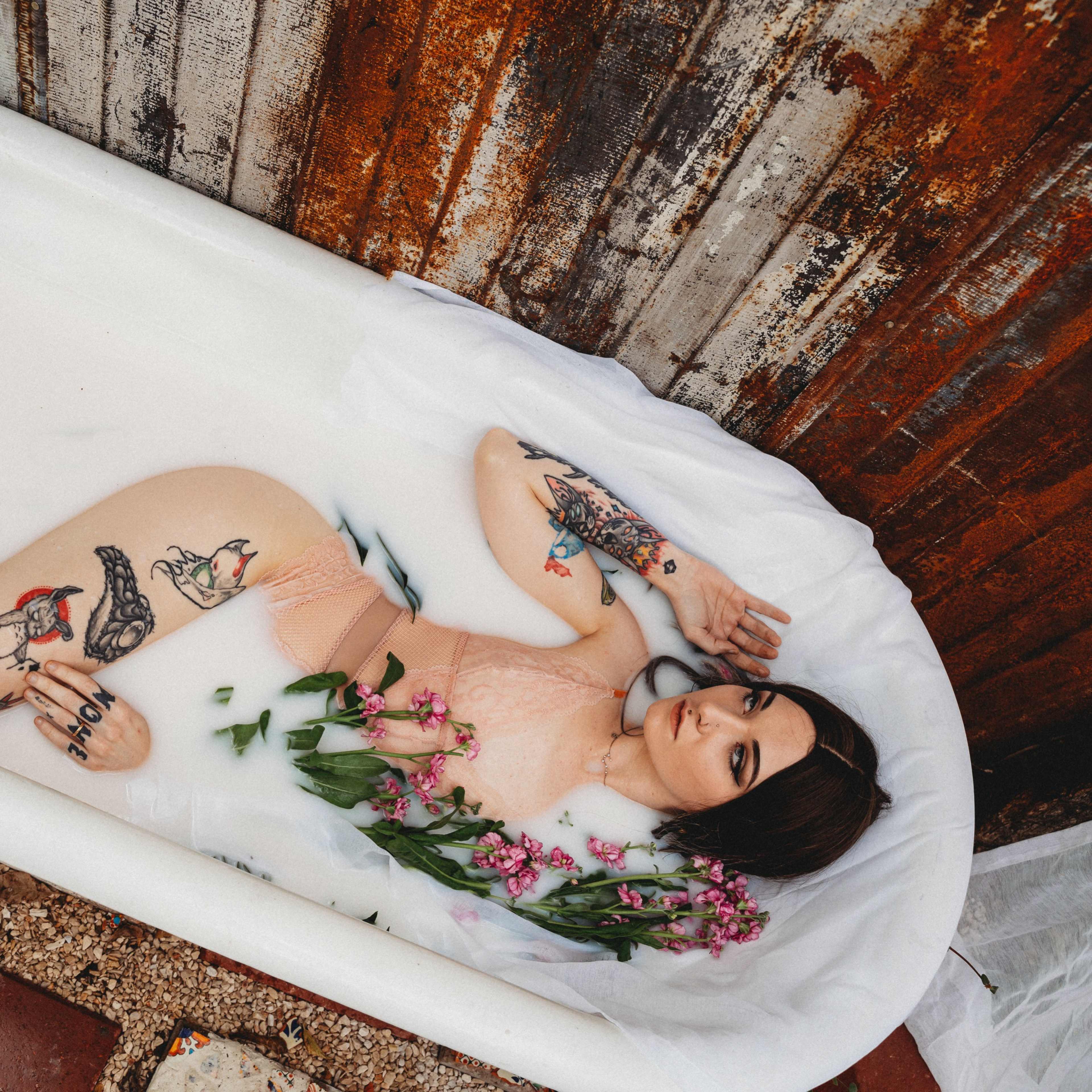 A person with various tattoos lies in a bathtub filled with milk and decorated with flowers, surrounded by rustic metal walls.