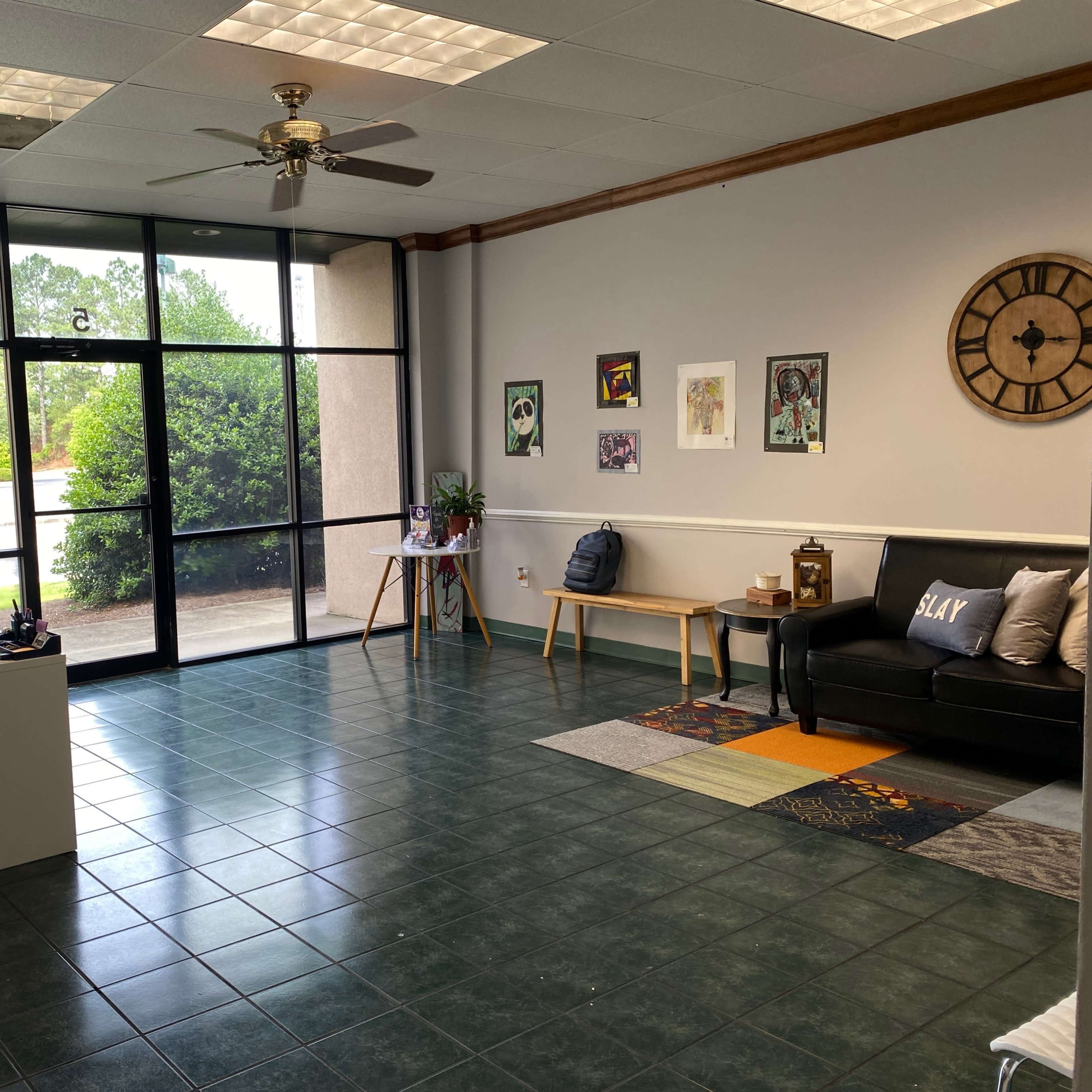An empty office space with a black sofa, a small desk, and large windows allowing natural light to enter.