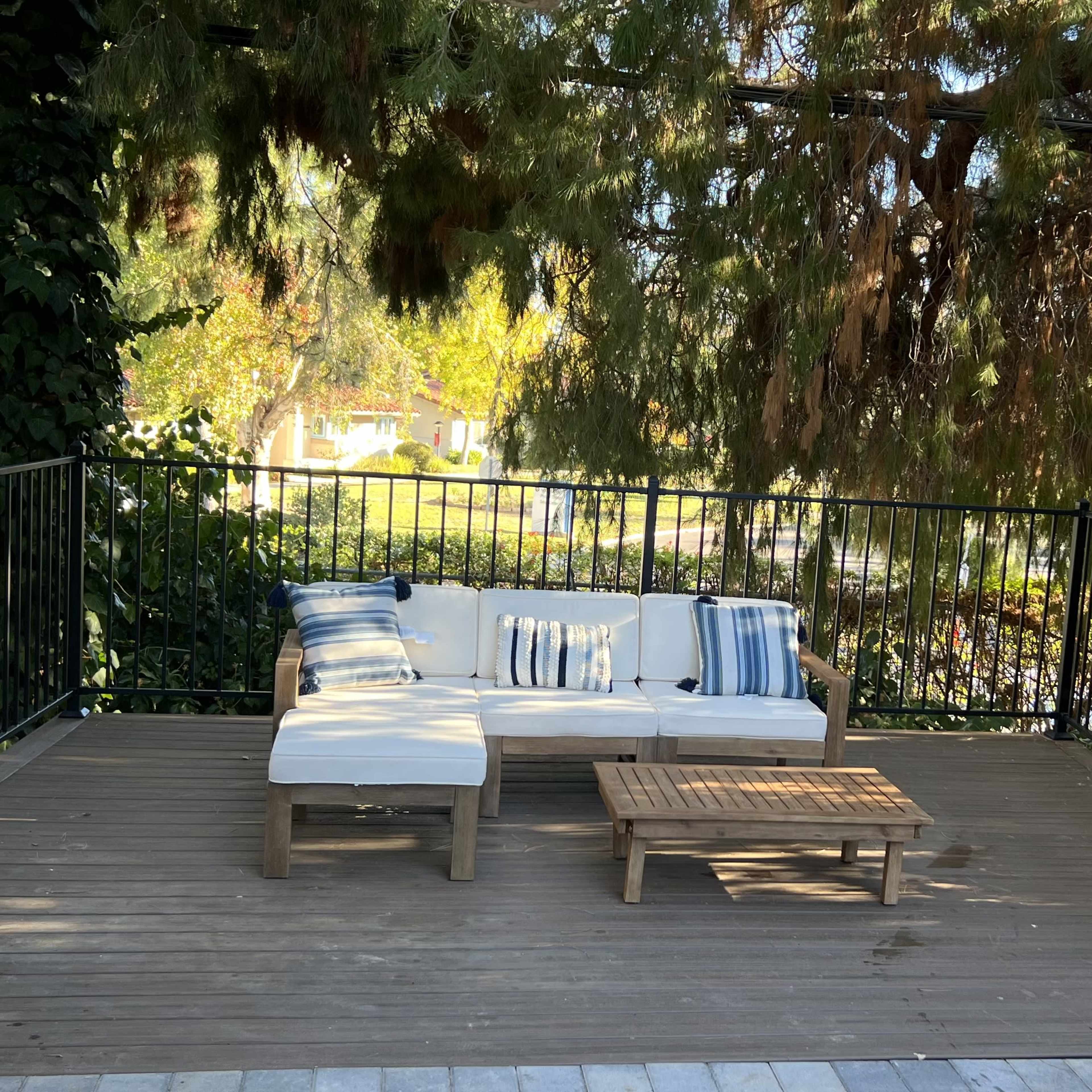 A wooden deck features a white sofa with blue accent pillows and a small coffee table, beside a patio heater and surrounded by greenery.