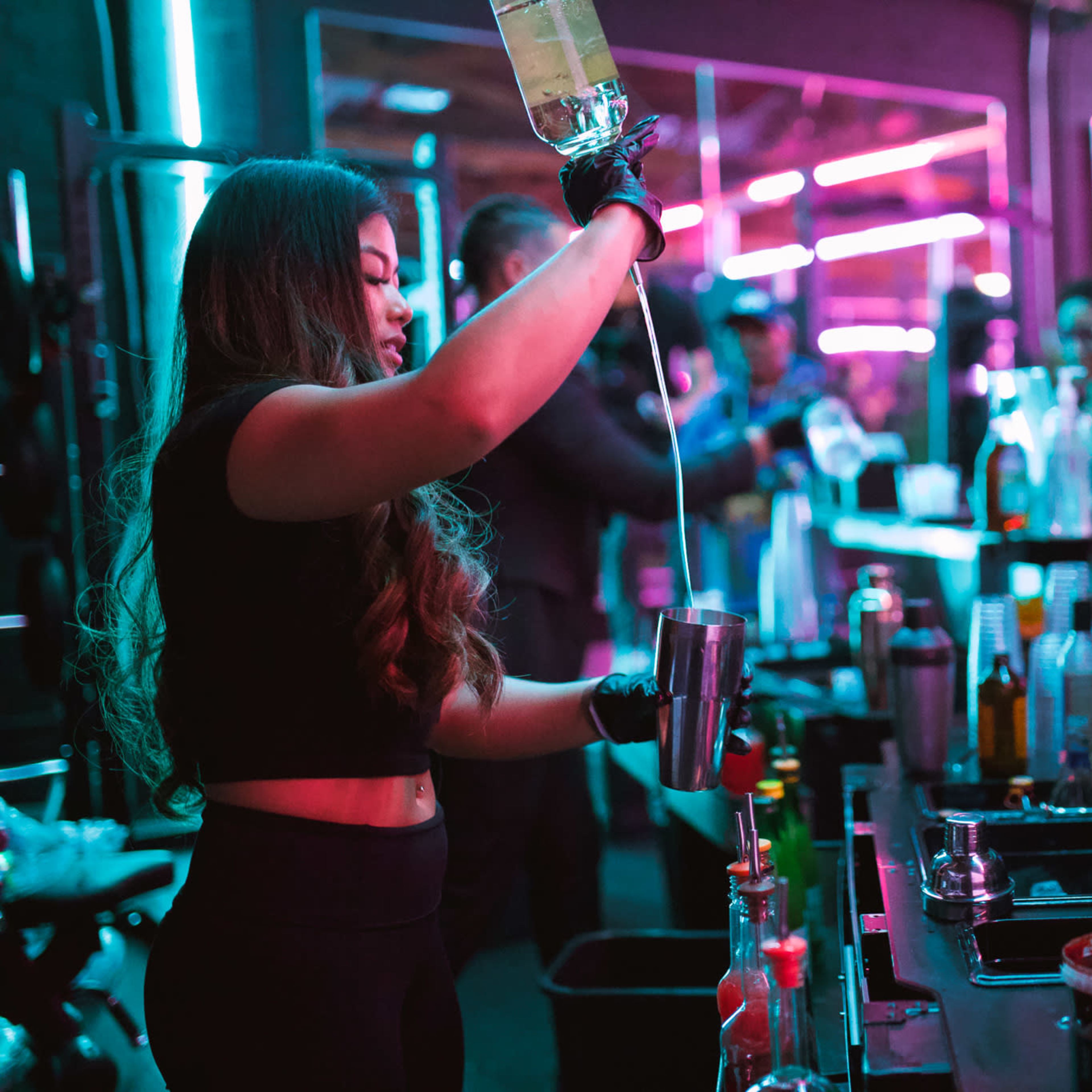 A bartender pours a drink from a bottle into a shaker at a vibrant bar illuminated by neon lights.