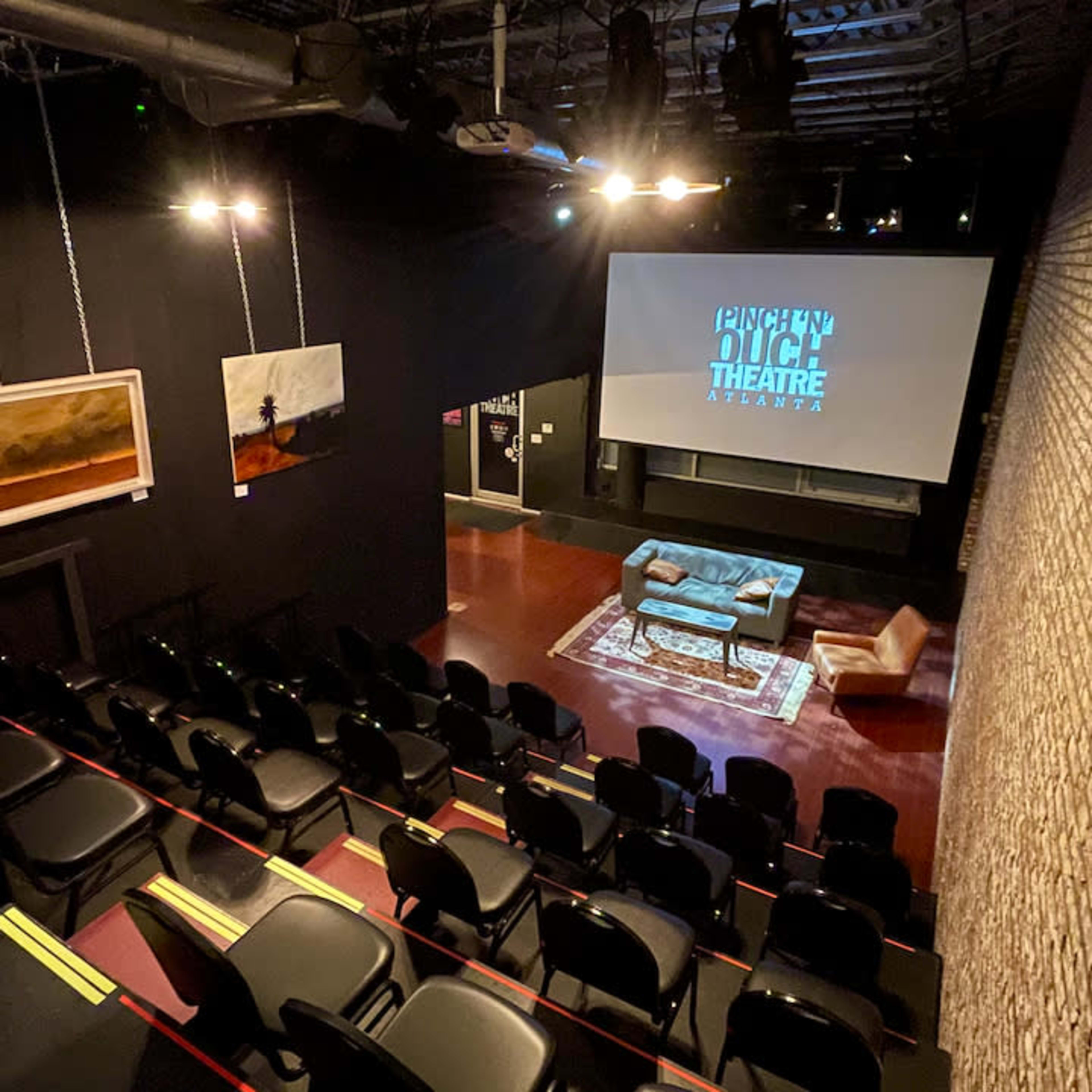 A small theater space with black seating and a large screen at the front, surrounded by brick walls and dim lighting.