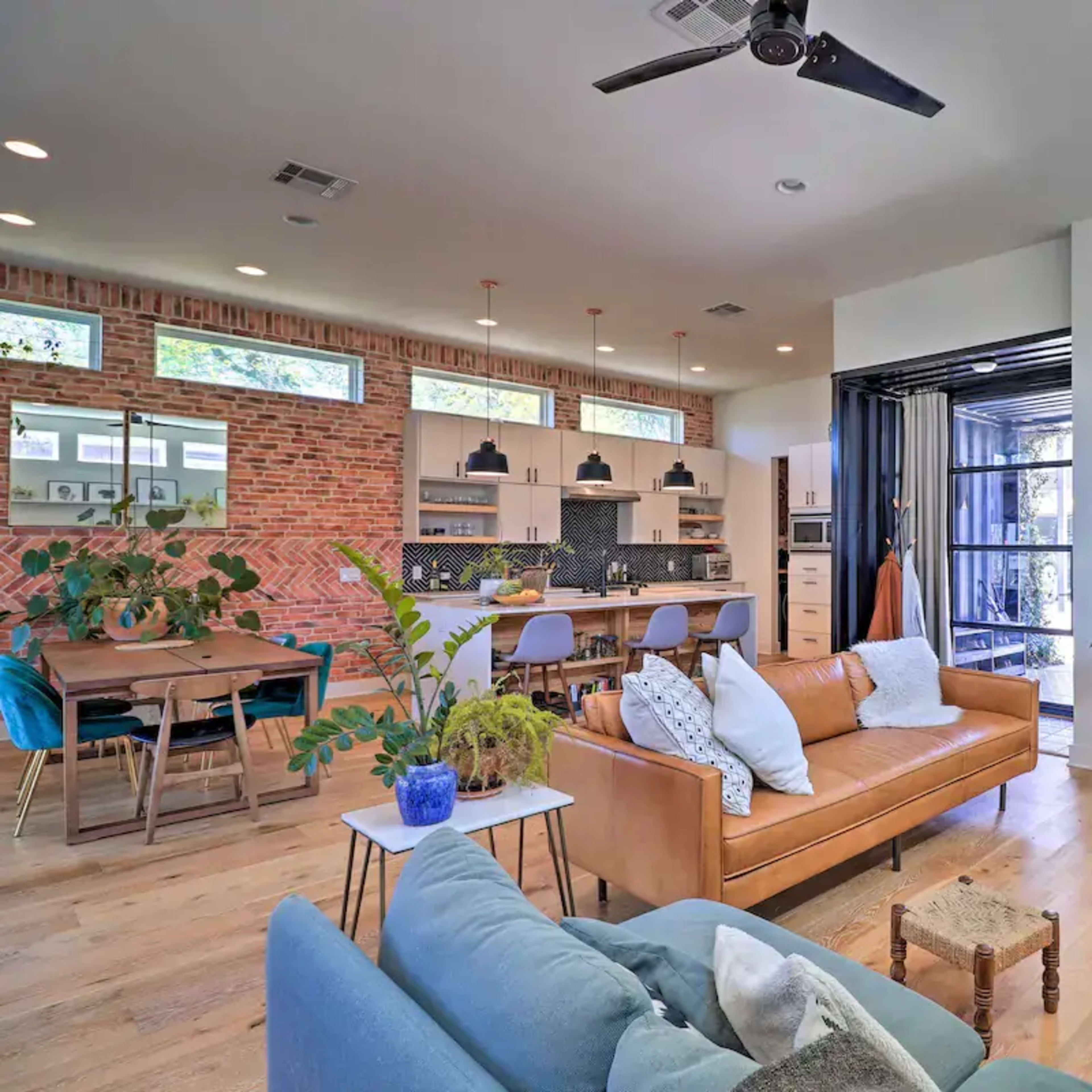 A modern open-concept living space featuring a living area with a leather sofa, a dining table, and a kitchen, all characterized by natural light and exposed brick walls.