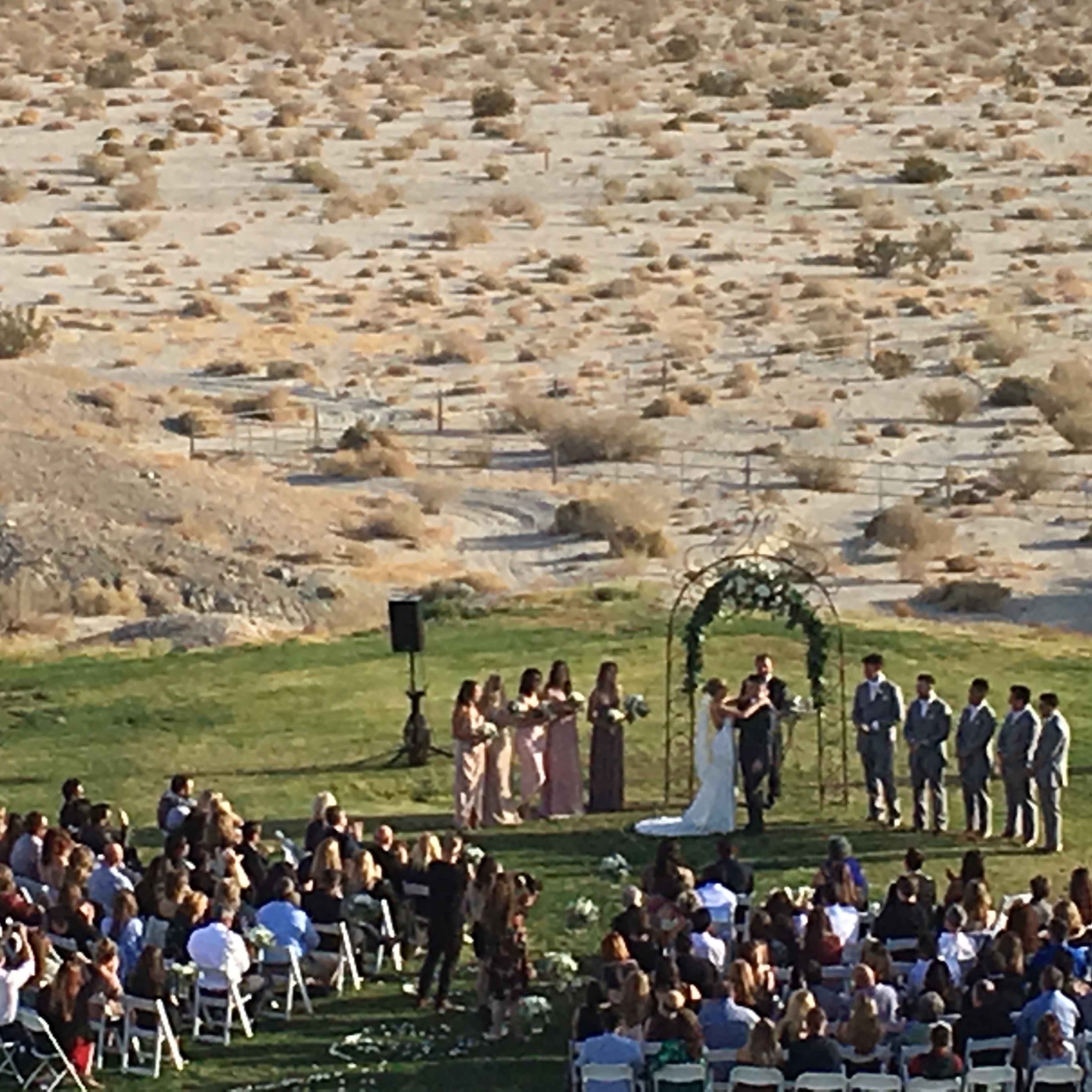 A couple stands under an arch during their wedding ceremony in a desert setting surrounded by guests seated in rows.
