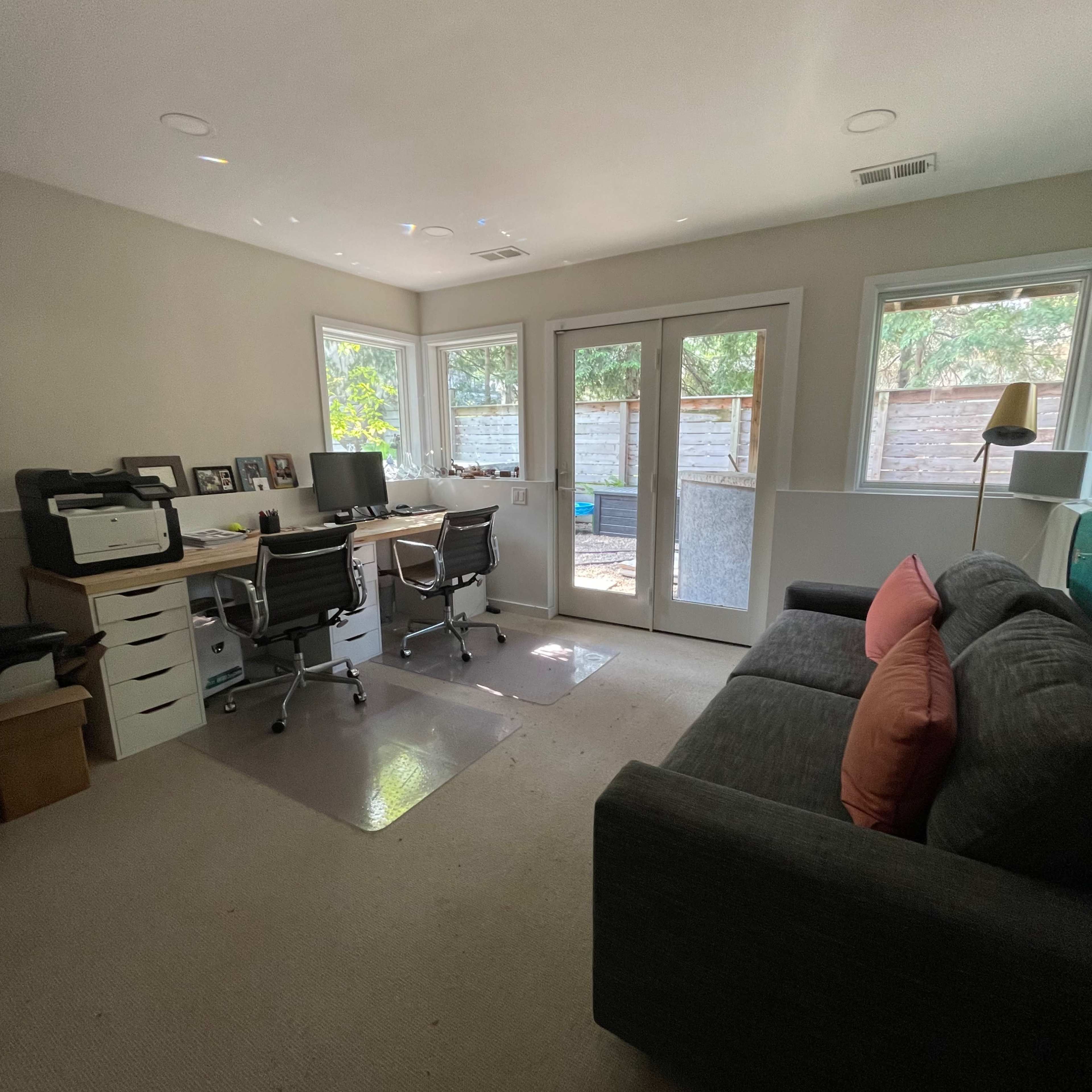 A home office with two desk setups, a printer, a gray sofa with colorful cushions, and large windows that provide natural light.