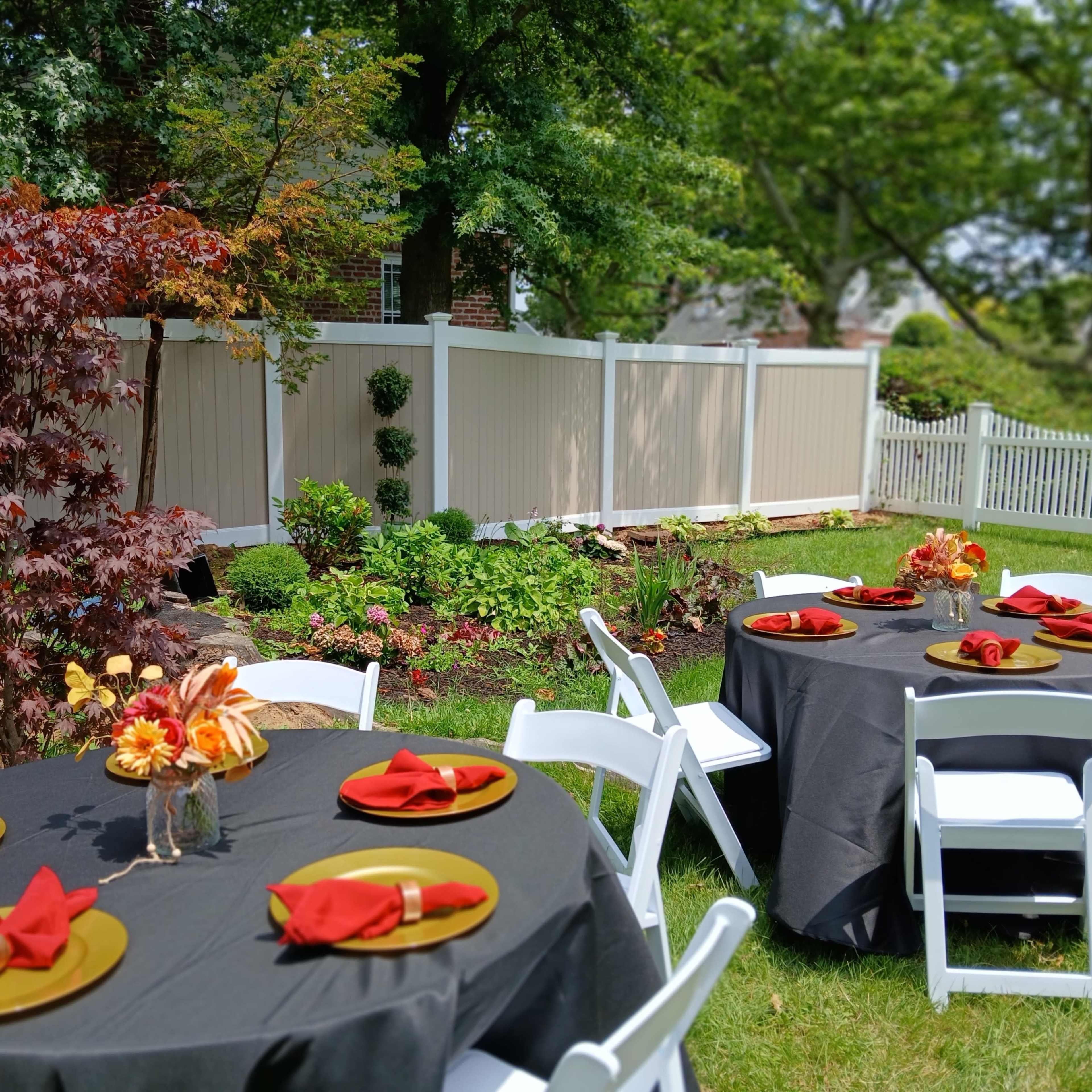 A backyard setting features round tables with black tablecloths, red napkins, and floral centerpieces, surrounded by greenery and a white picket fence.