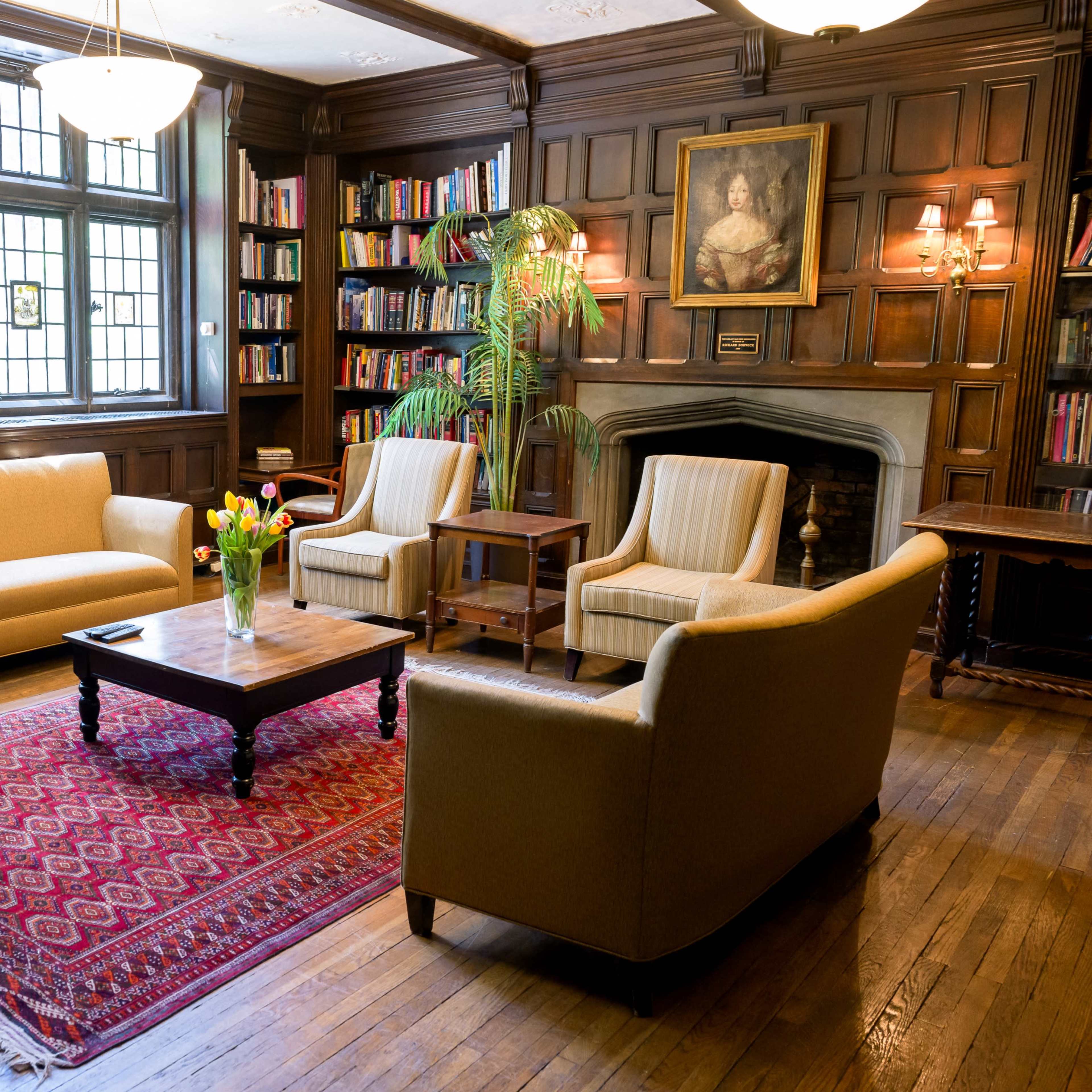A cozy living room with wooden paneling, featuring a sofa, two armchairs, a coffee table, and a colorful area rug, alongside a bookshelf and a fireplace.