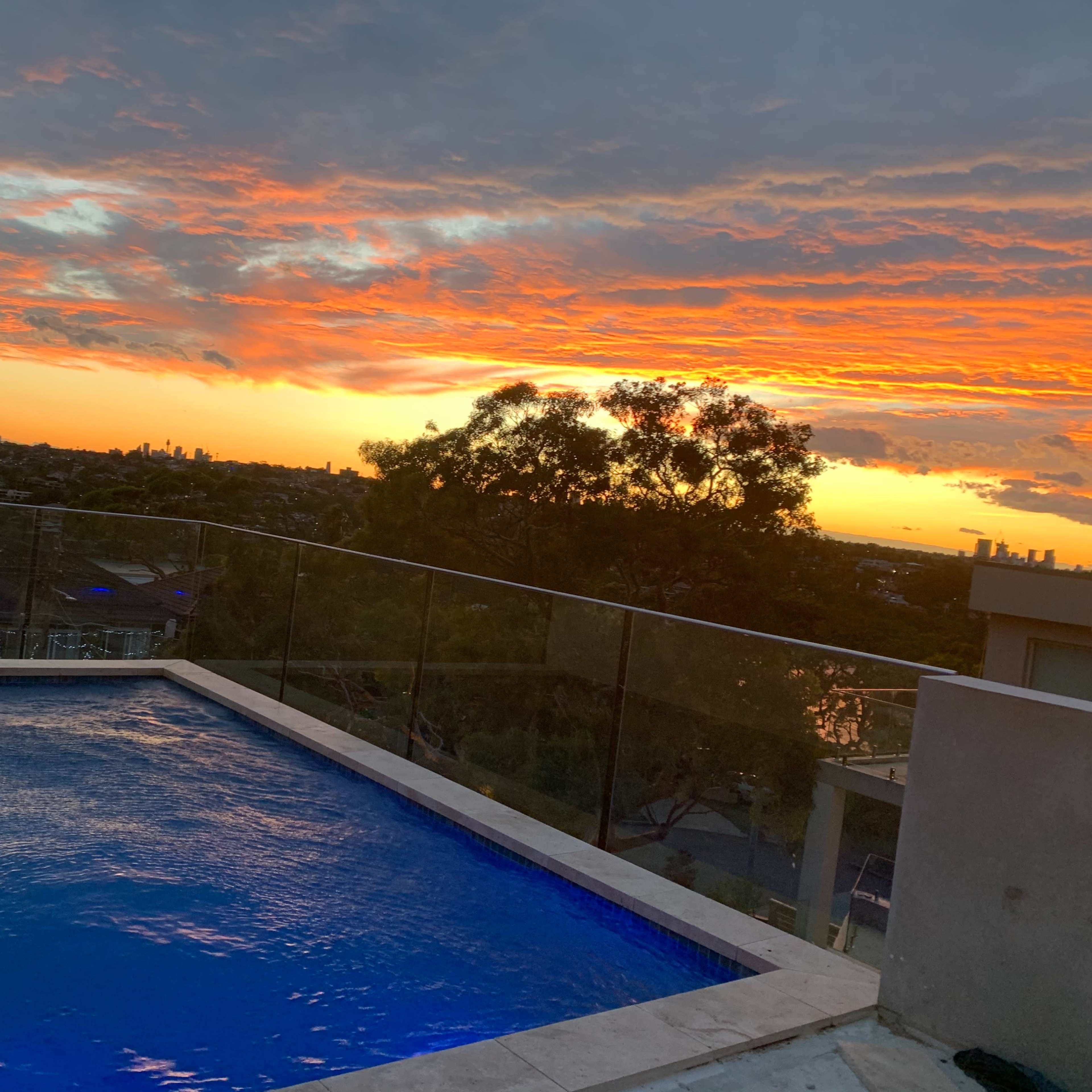 A rooftop swimming pool overlooking a city skyline at sunset, with vibrant orange and purple clouds in the sky.