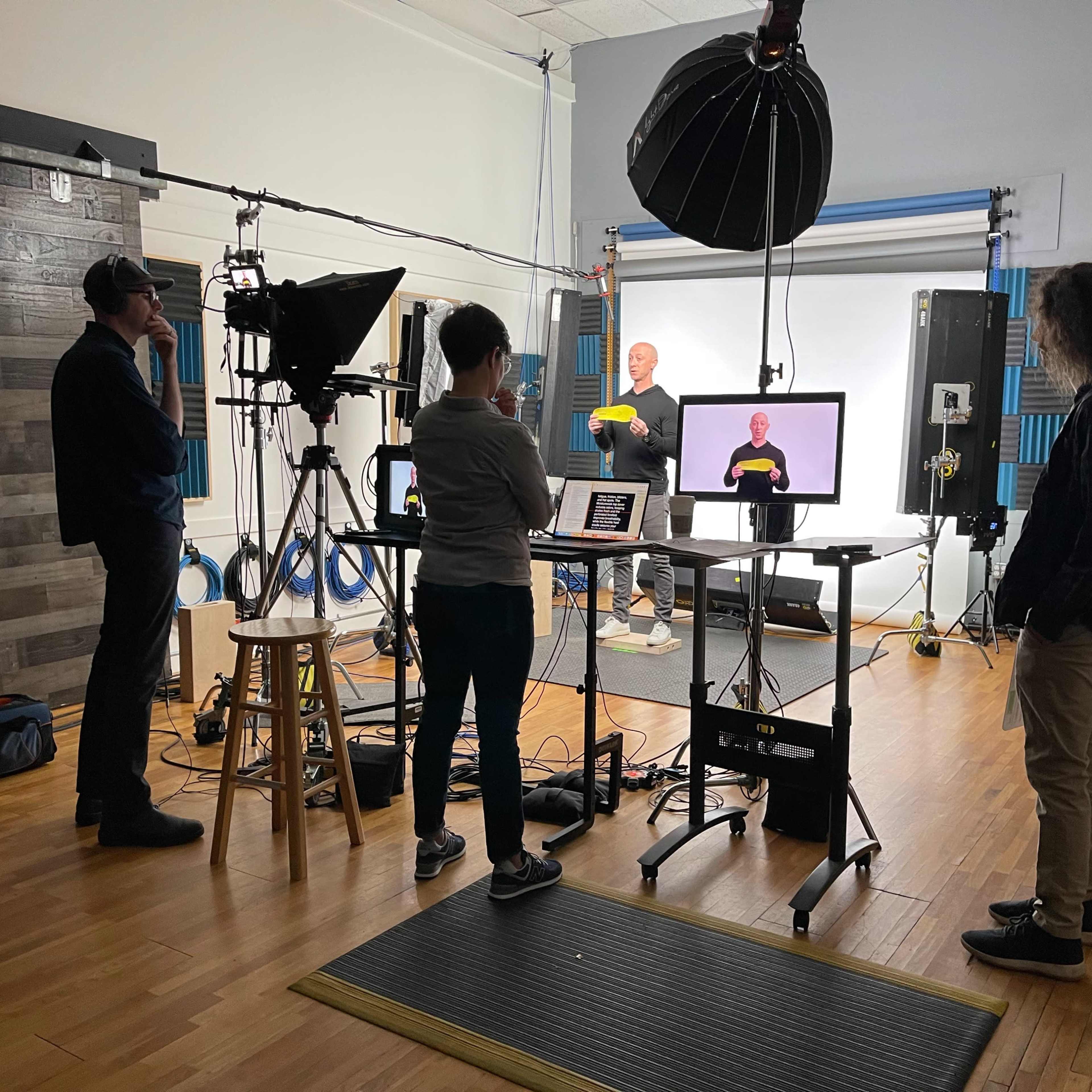 A group of four people is gathered in a studio setting, with one person in front of a screen presenting while the others observe and operate equipment.