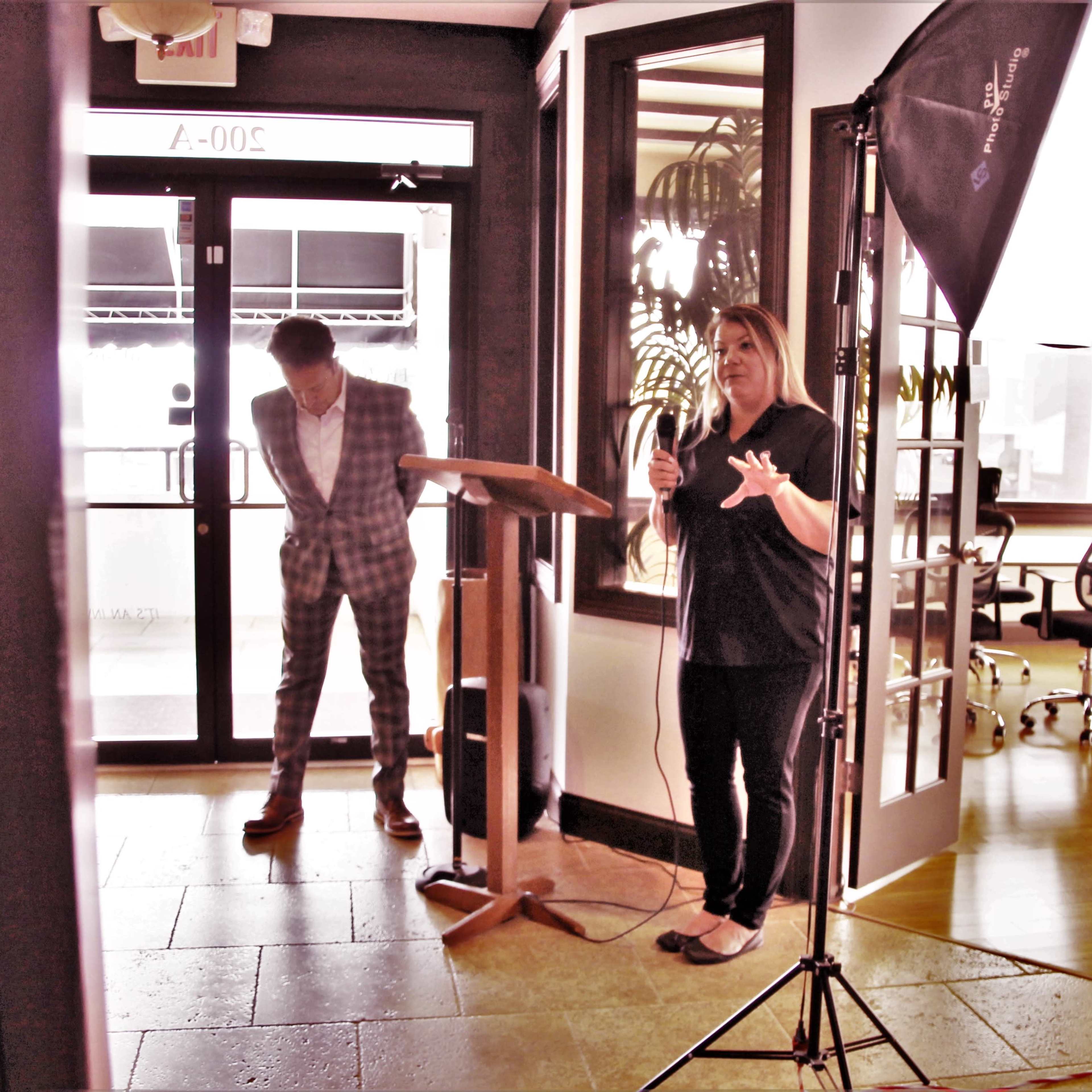 A woman speaks at a podium in a modern office space while a man in a checked suit stands nearby.