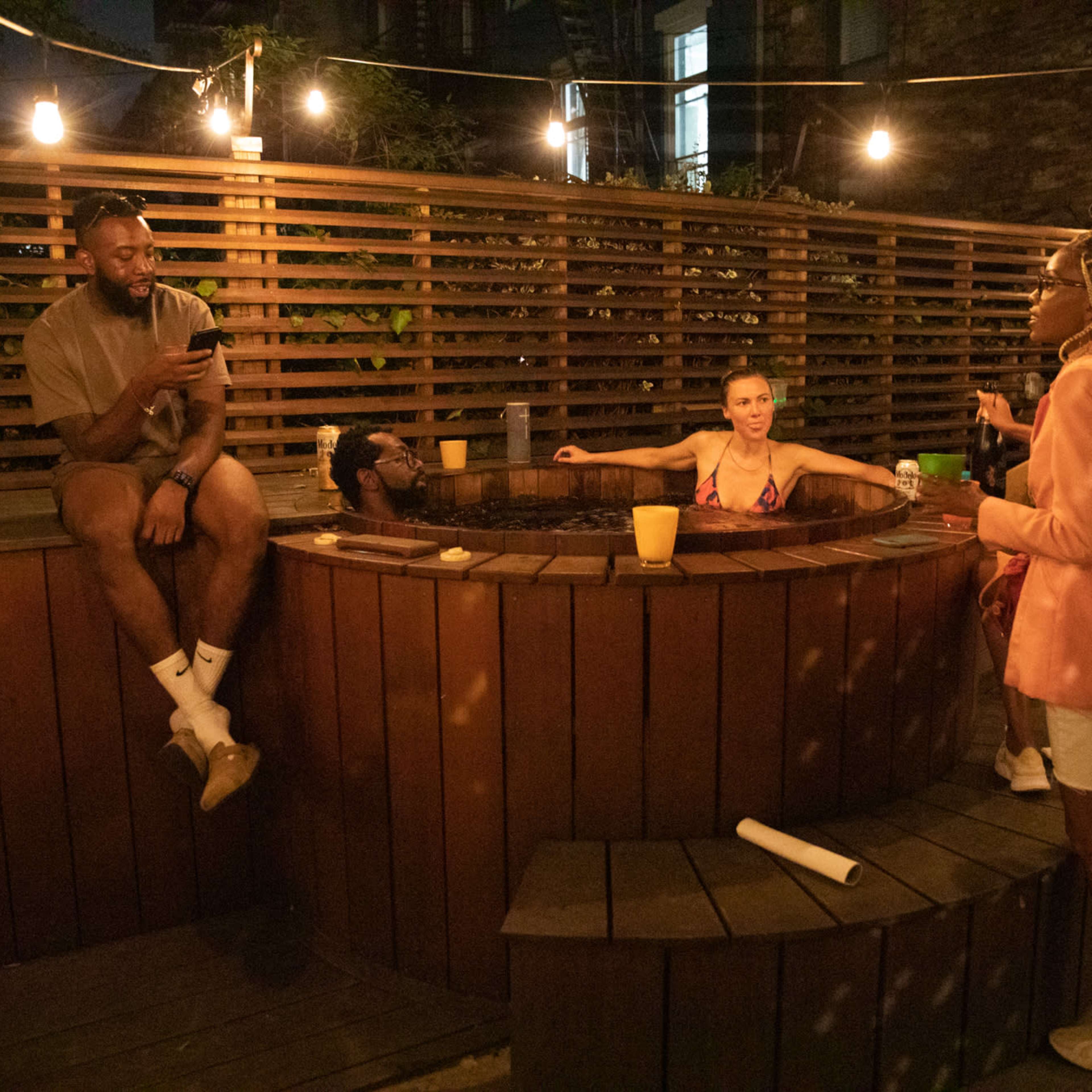 A group of four people socializes around a wooden hot tub in a dimly lit outdoor space, with string lights overhead.