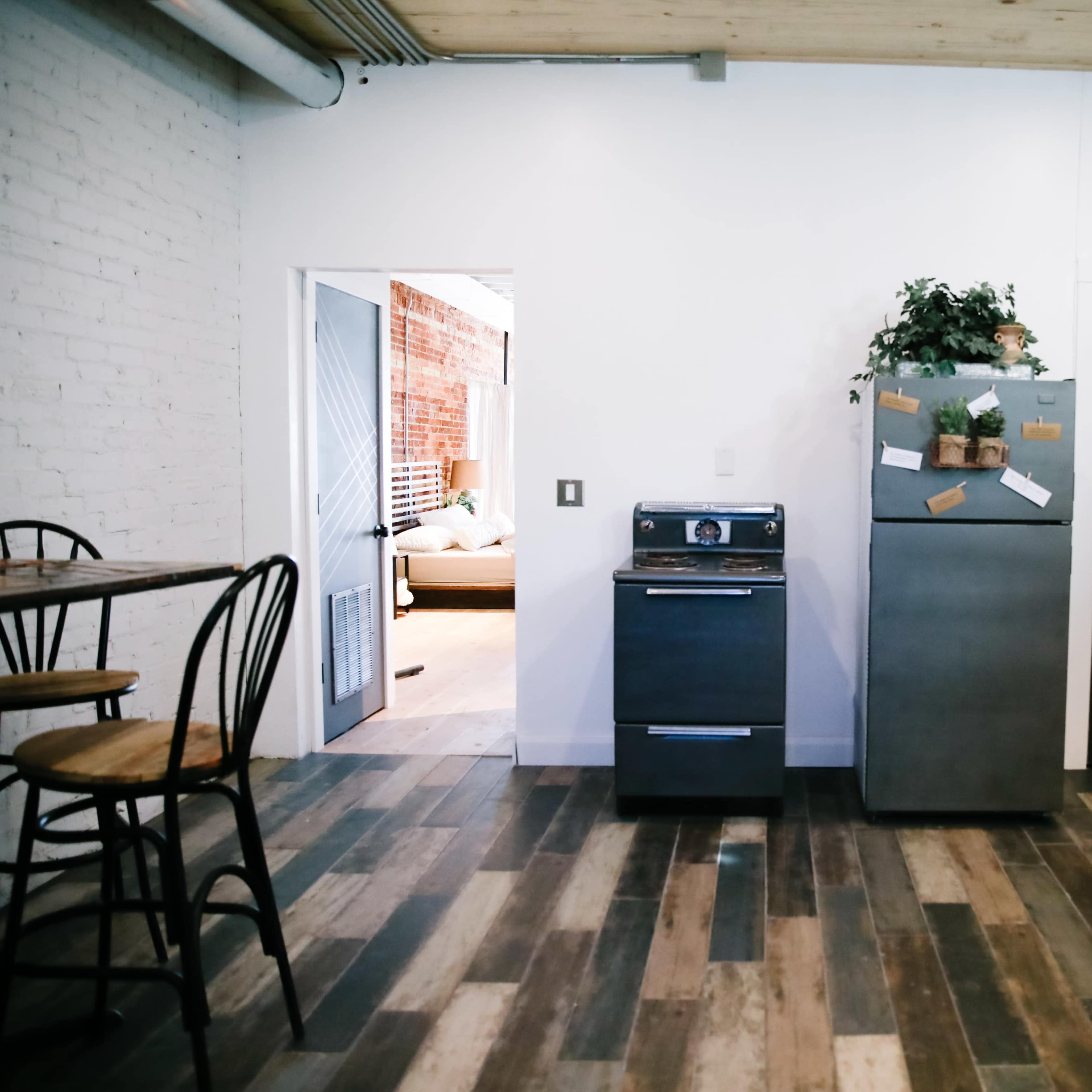 A modern kitchen area with a refrigerator and an oven, featuring a dining table with stools and a view of an adjacent room through an open door.