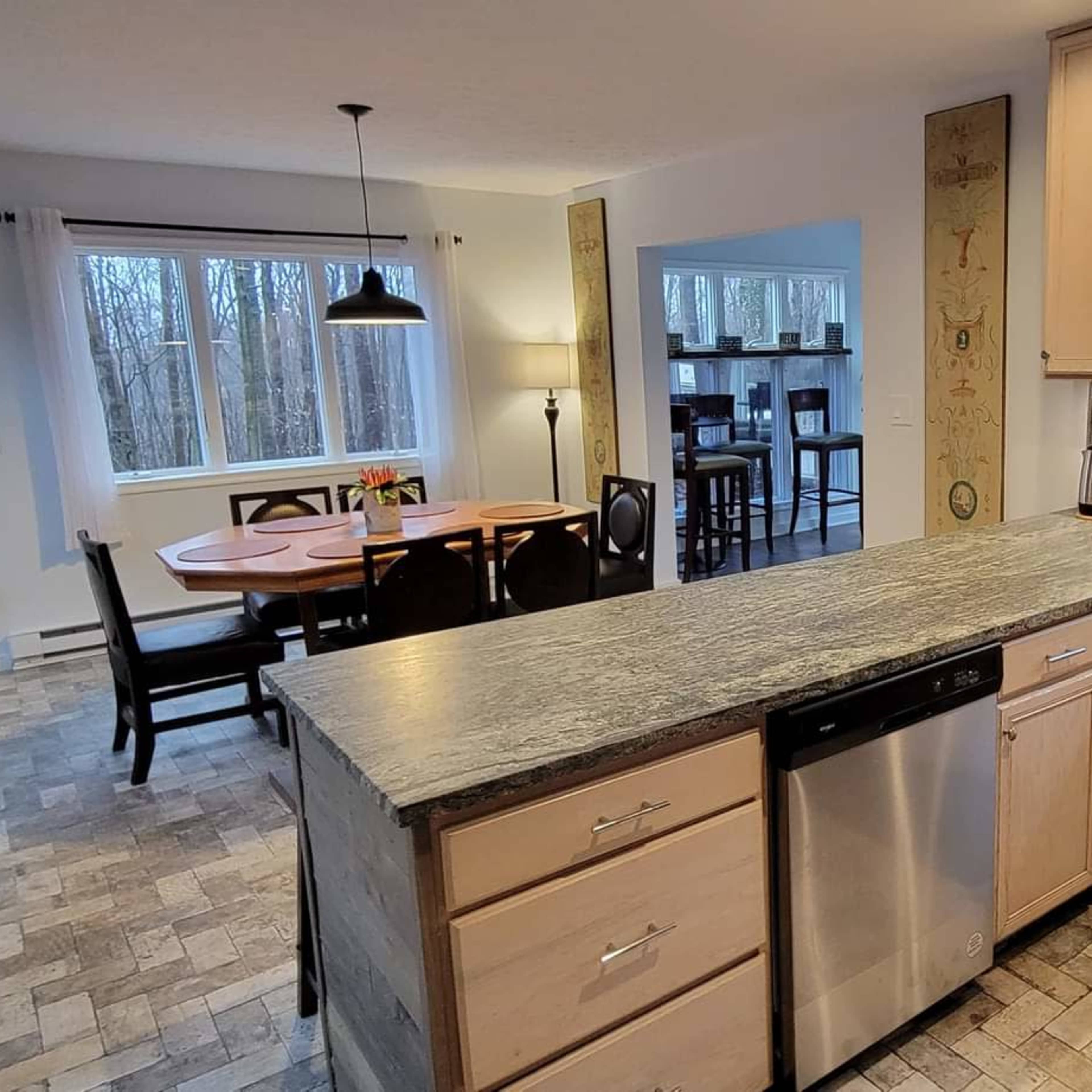 A modern kitchen with light wood cabinetry, a stainless steel dishwasher, and a view of an adjoining dining area with a round table and chairs.