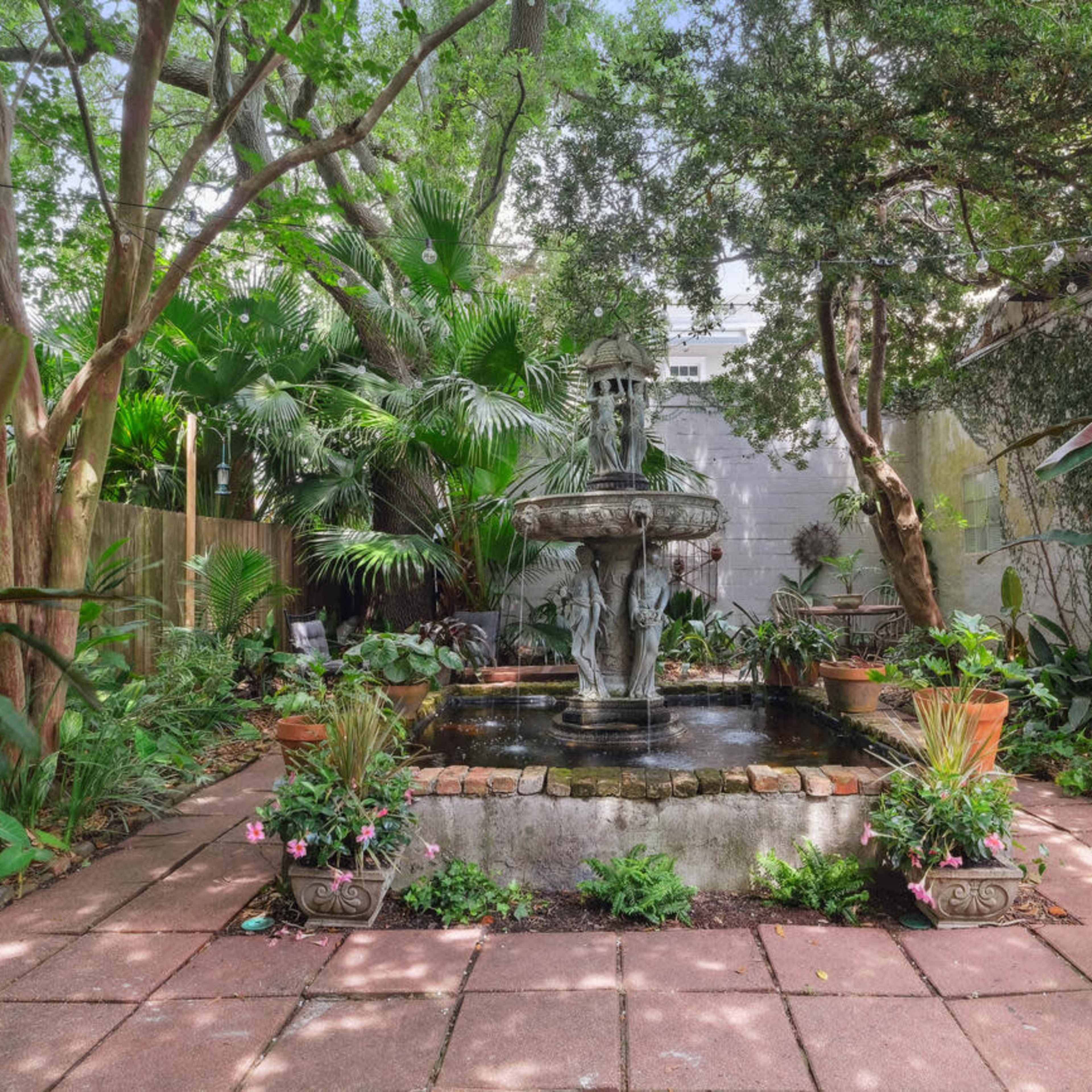 A stone fountain surrounded by lush greenery and potted plants is situated in a brick-paved courtyard.