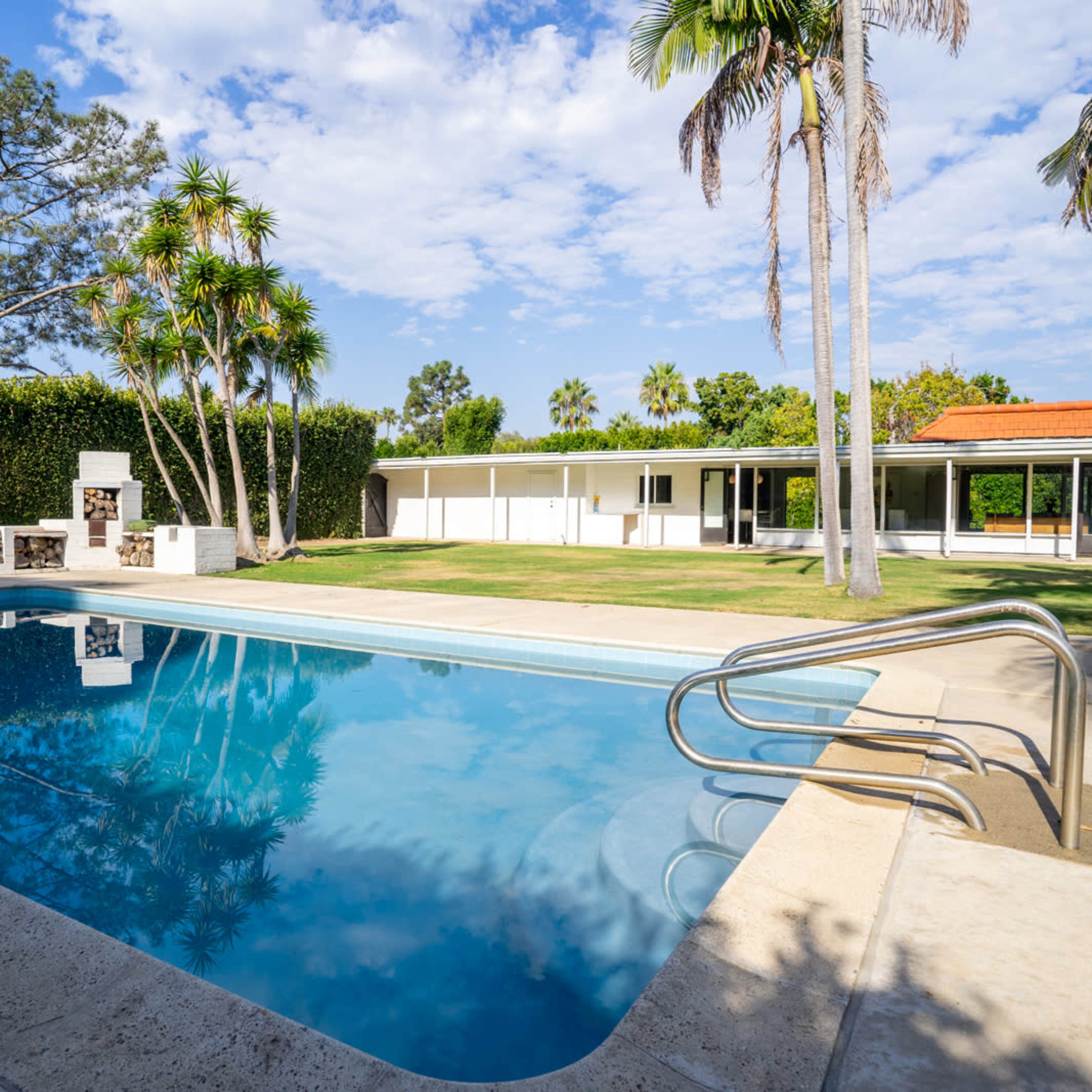 A clear swimming pool surrounded by a manicured lawn and palm trees, with a house and outdoor barbecue area visible in the background.