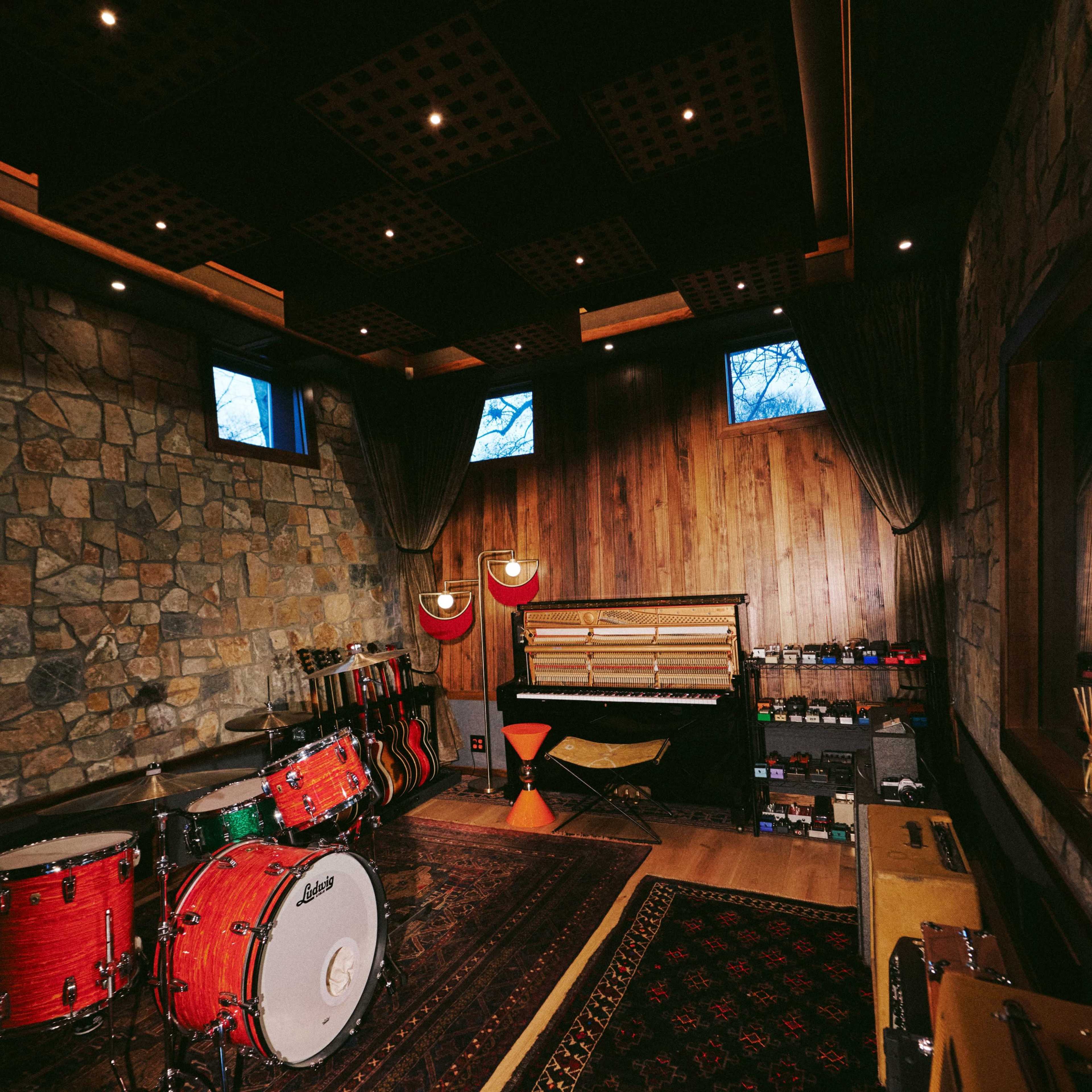 A music studio features a red drum set, a piano, and shelves with various instruments against a stone and wood backdrop.