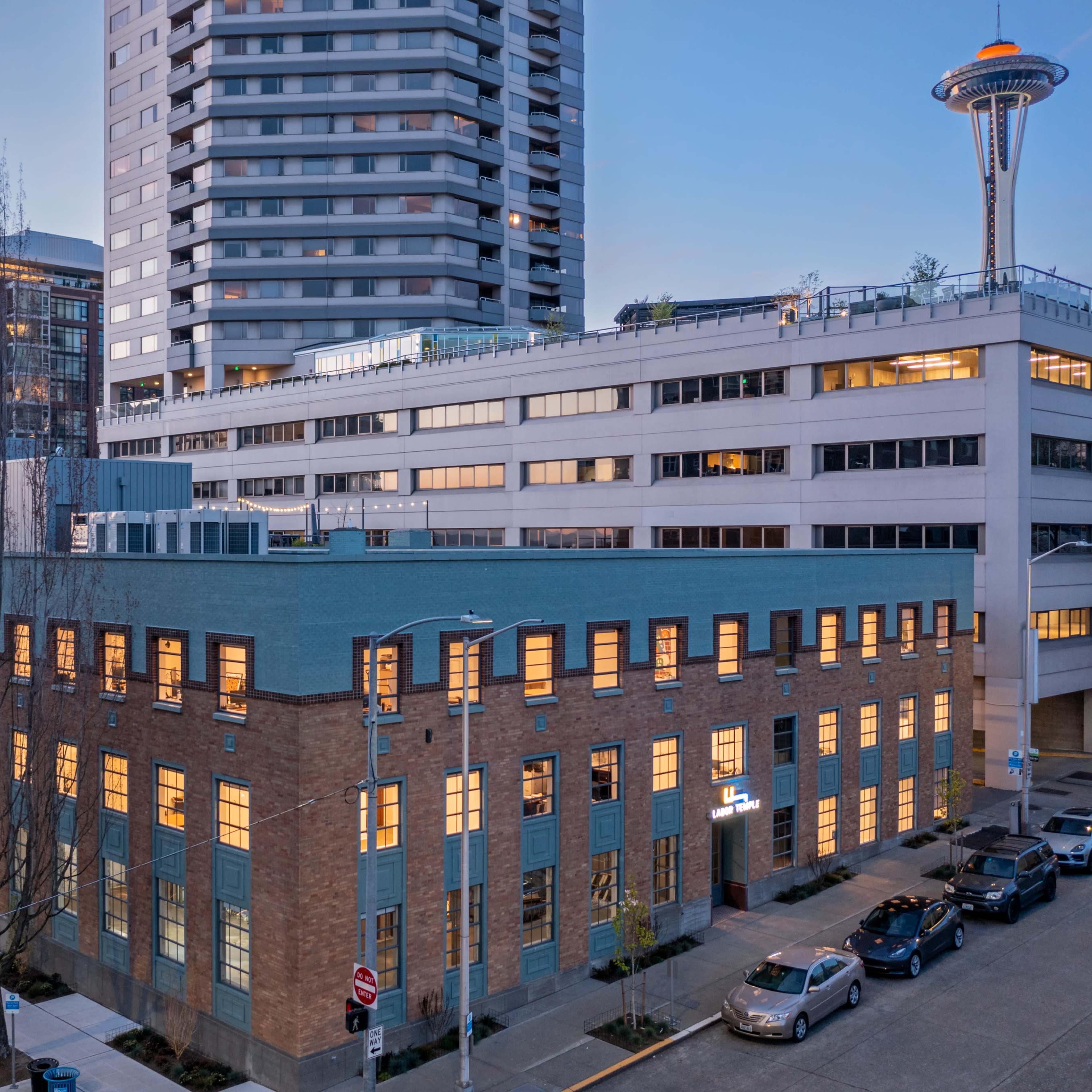 A modern high-rise building alongside a low-rise brick structure, with the Space Needle visible in the background.