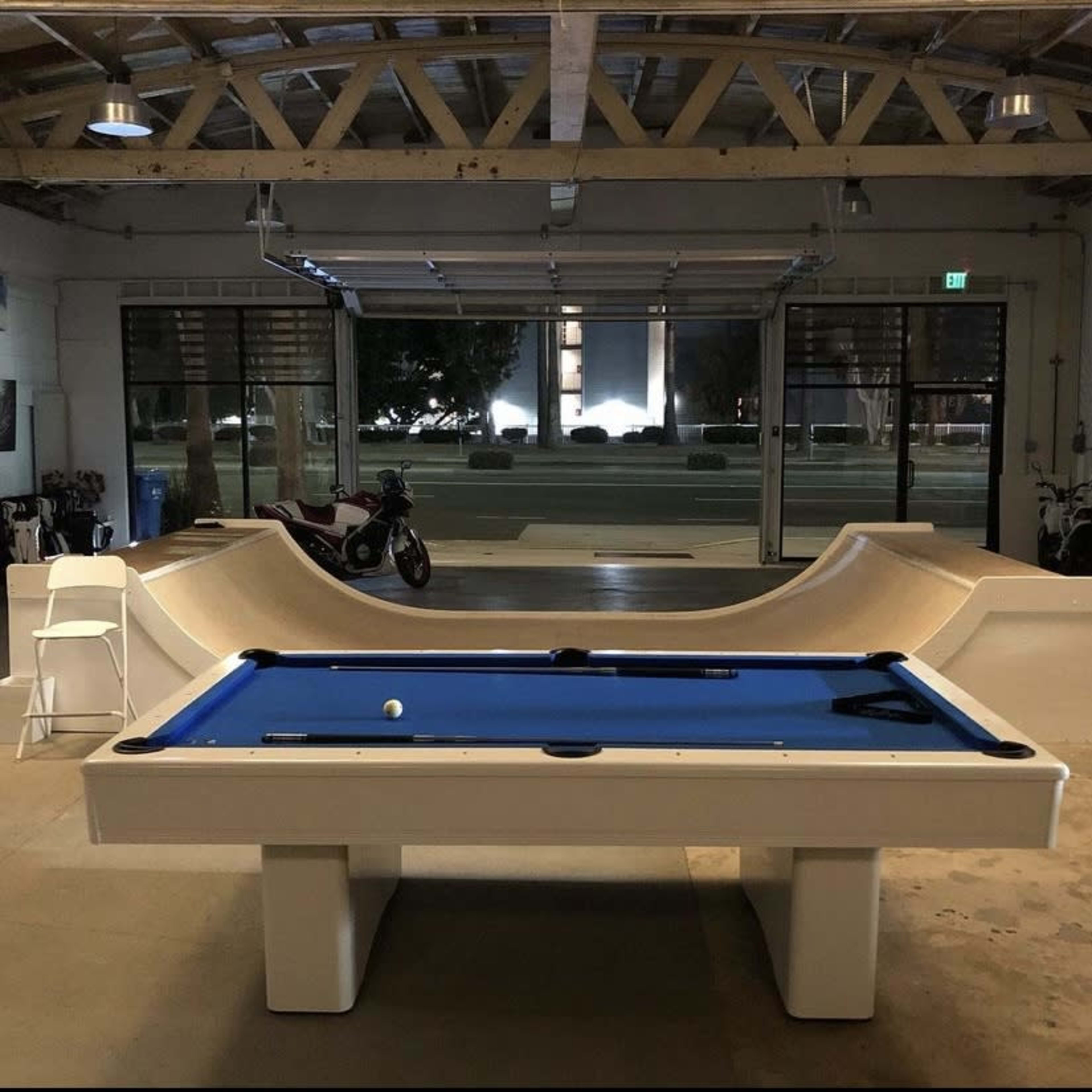 A blue pool table is positioned in an industrial garage space with a motorcycle and a skateboard ramp visible in the background.
