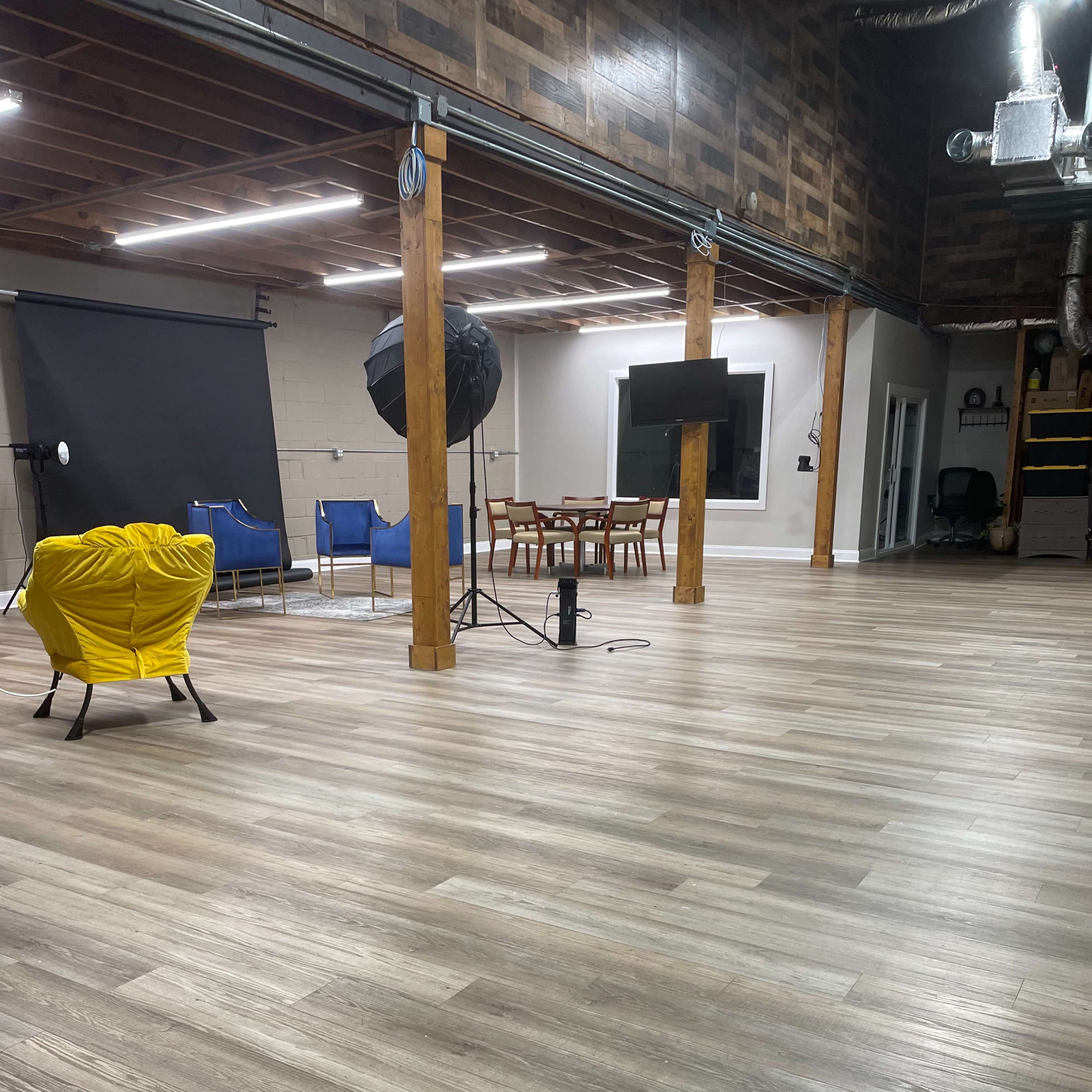 An empty studio space with wooden flooring, equipped with lighting equipment, chairs, a dining table, and a television mounted on the wall.