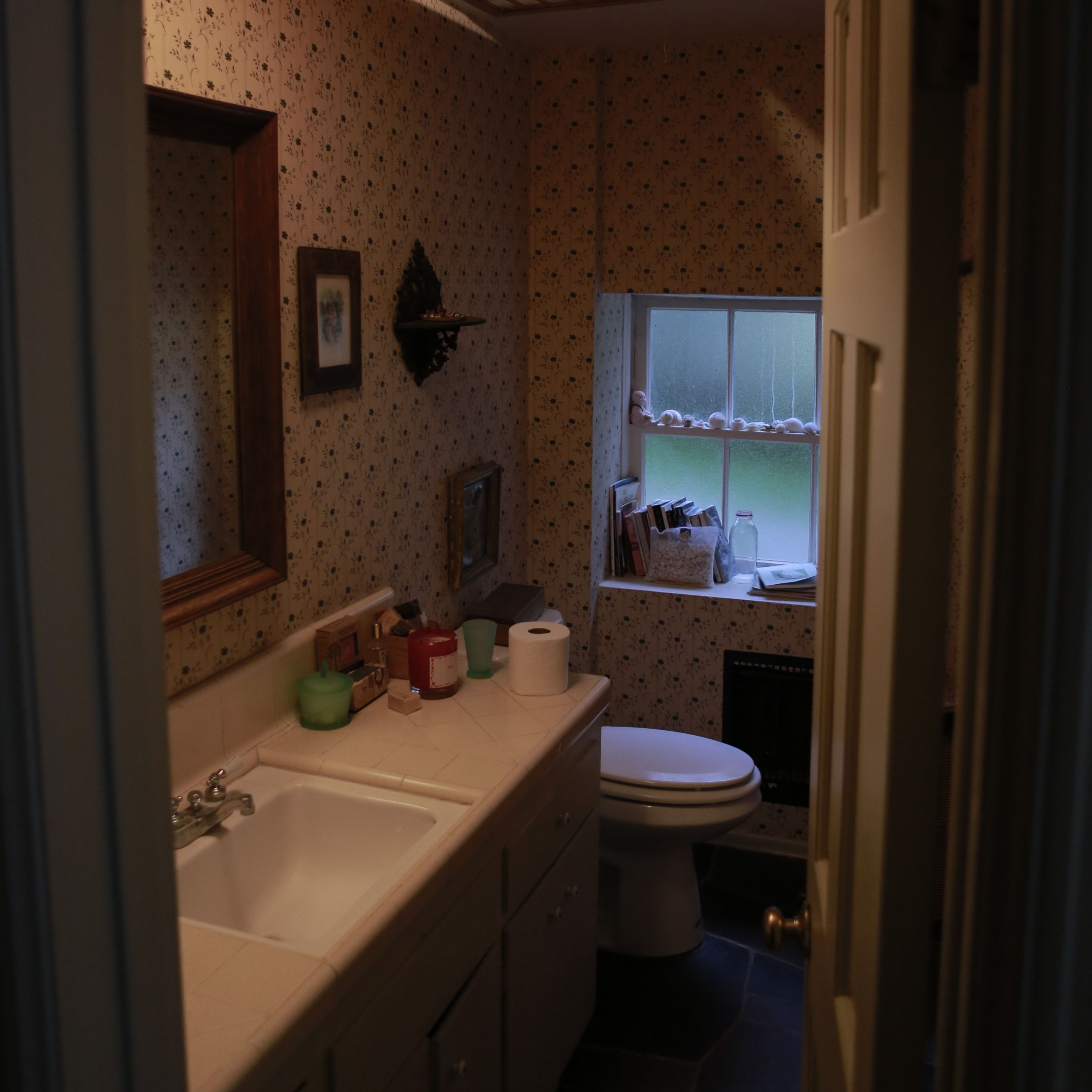 A small bathroom with a sink, toilet, and window, featuring floral wallpaper and a mirror.