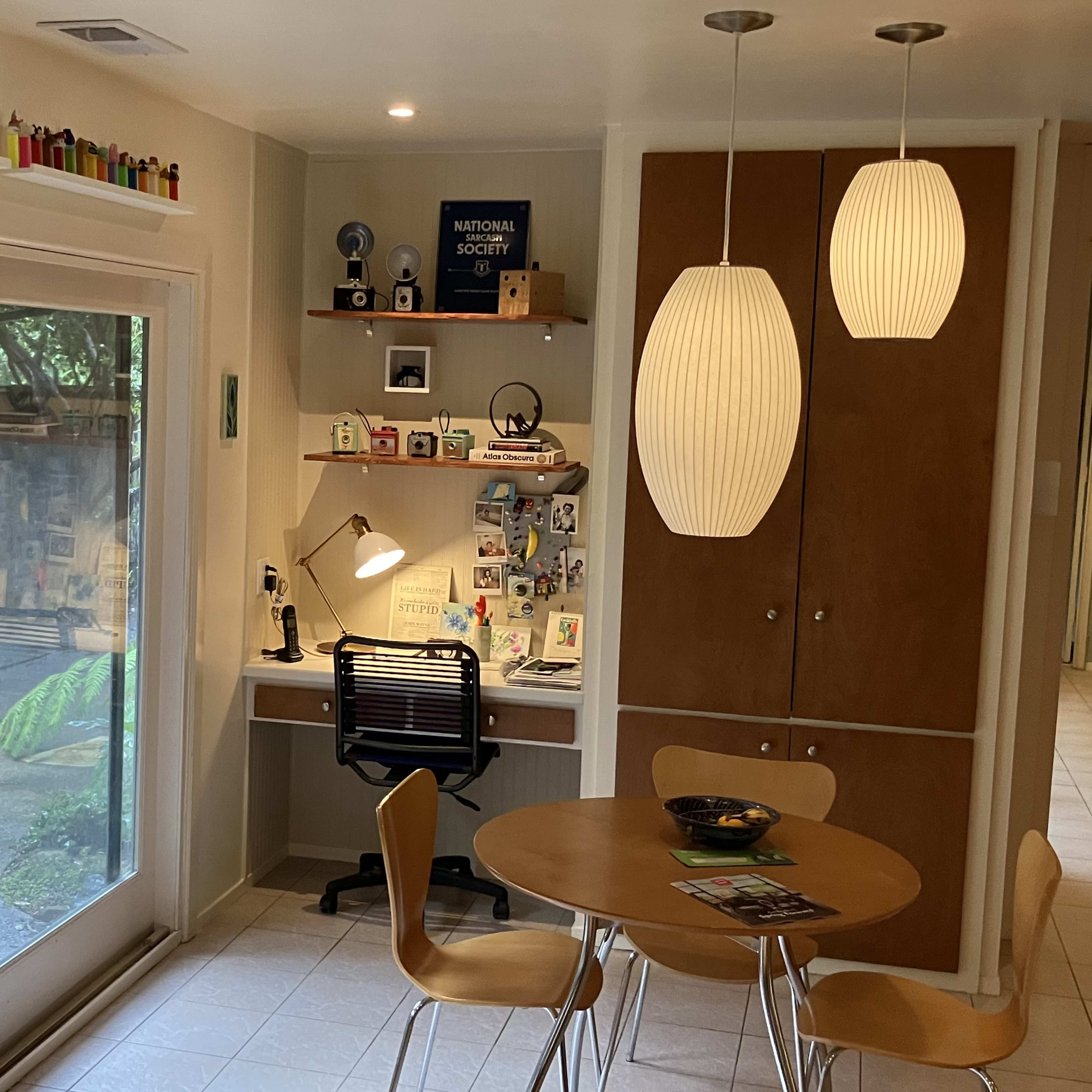 A modern kitchen area features a round wooden table, a desk with office supplies, and large sliding glass doors that open to an outdoor space.