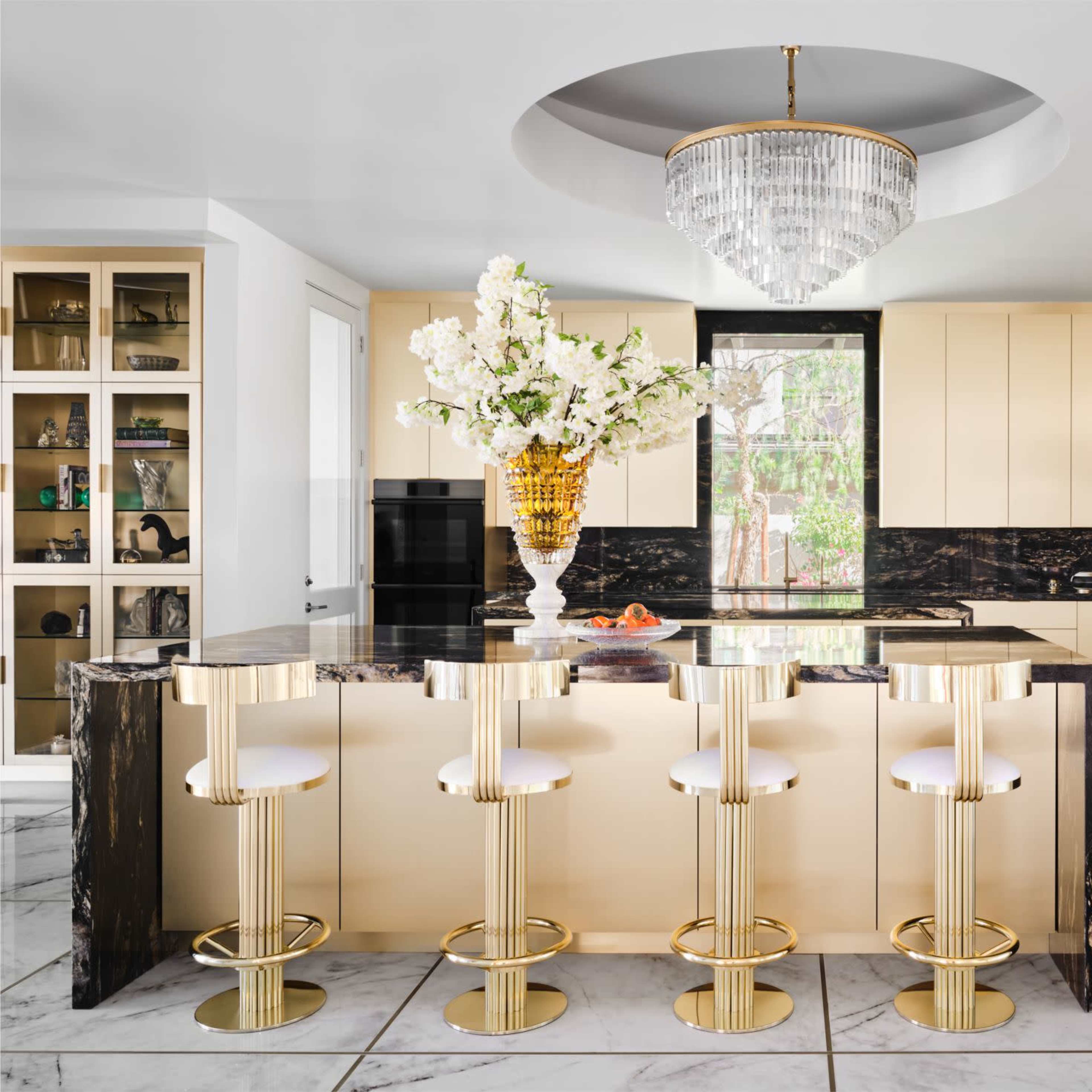A modern kitchen features a central marble island with gold-accented stools, light wood cabinetry, and a large chandelier overhead.
