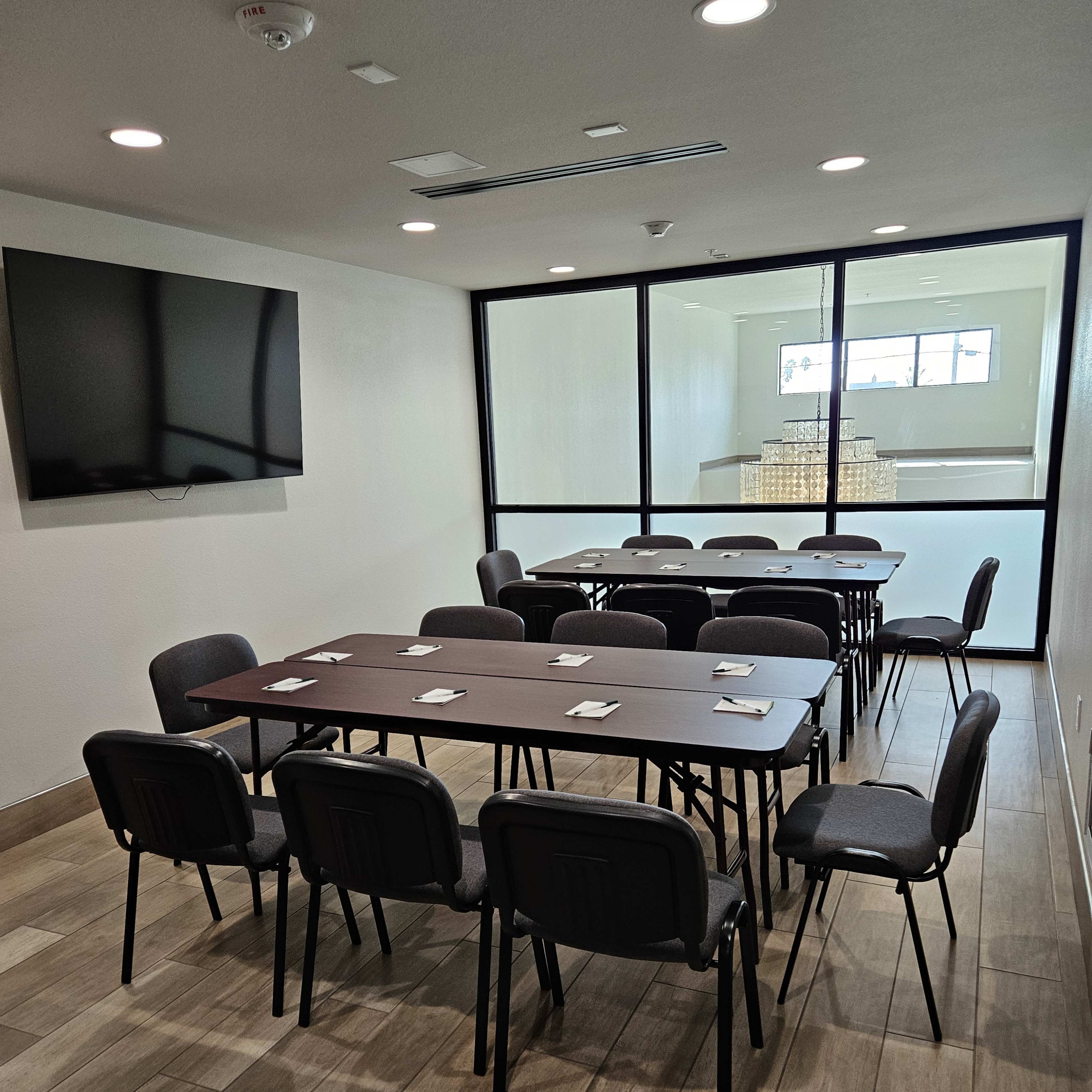 A conference room features a long table surrounded by chairs, with a television mounted on the wall and a glass partition leading to another room.