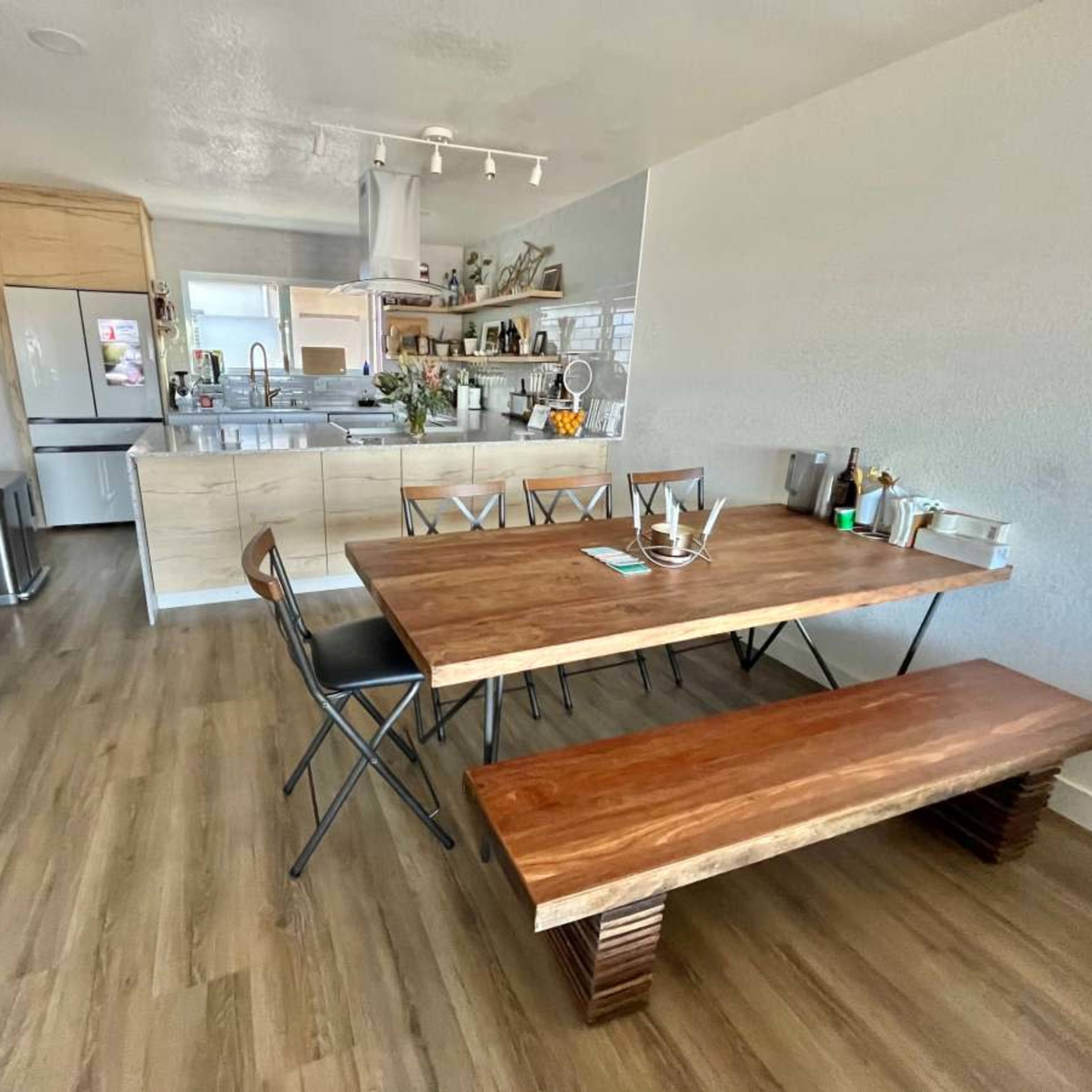 A modern dining area features a wooden table and bench, with black folding chairs, adjacent to an open kitchen with light cabinetry and appliances.